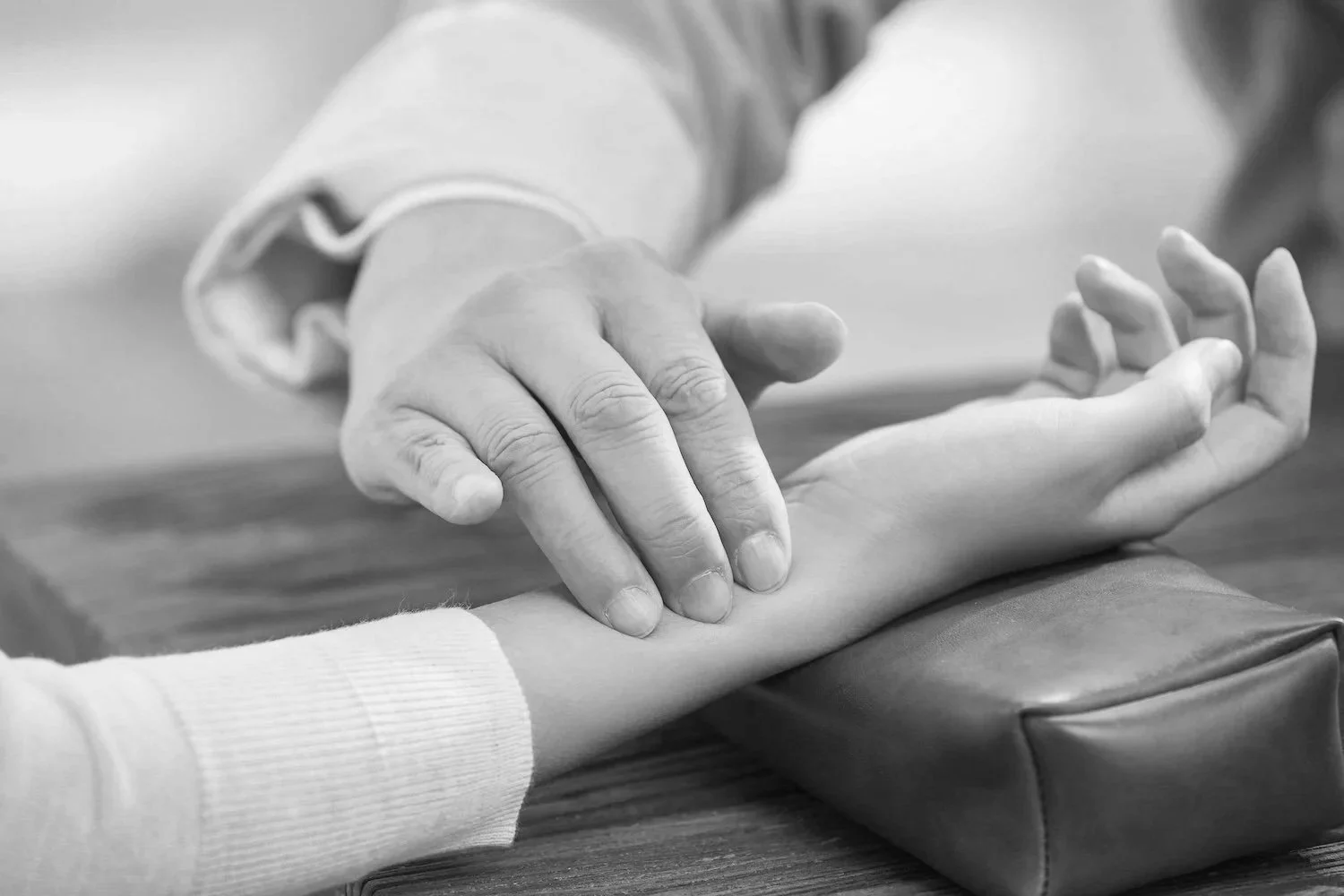 An acupuncturist checking a patient's wrist for a pulse, with a blood pressure cuff on the patient's arm, in black and white.