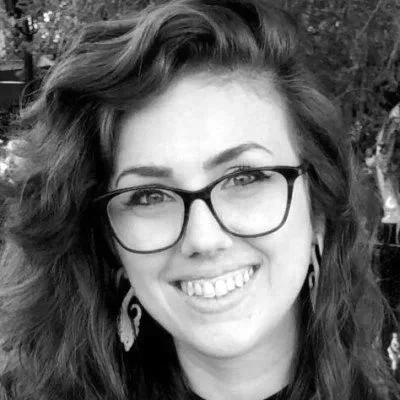 A woman with curly hair, wearing glasses and earrings, smiling outdoors.