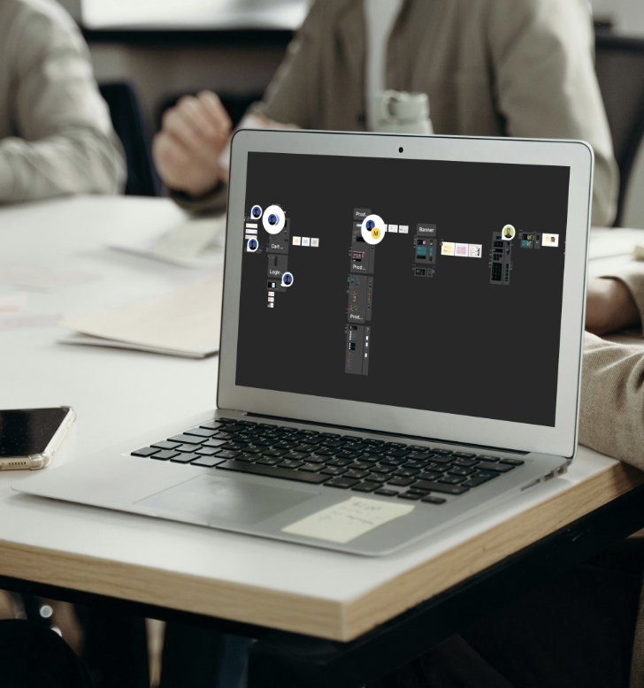 A laptop with a digital workspace or flowchart displayed on the screen, placed on a table in a meeting room where several people are sitting.