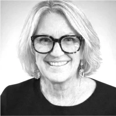 A black-and-white portrait of a woman with shoulder-length hair, wearing glasses, earrings, and a dark top, smiling at the camera.