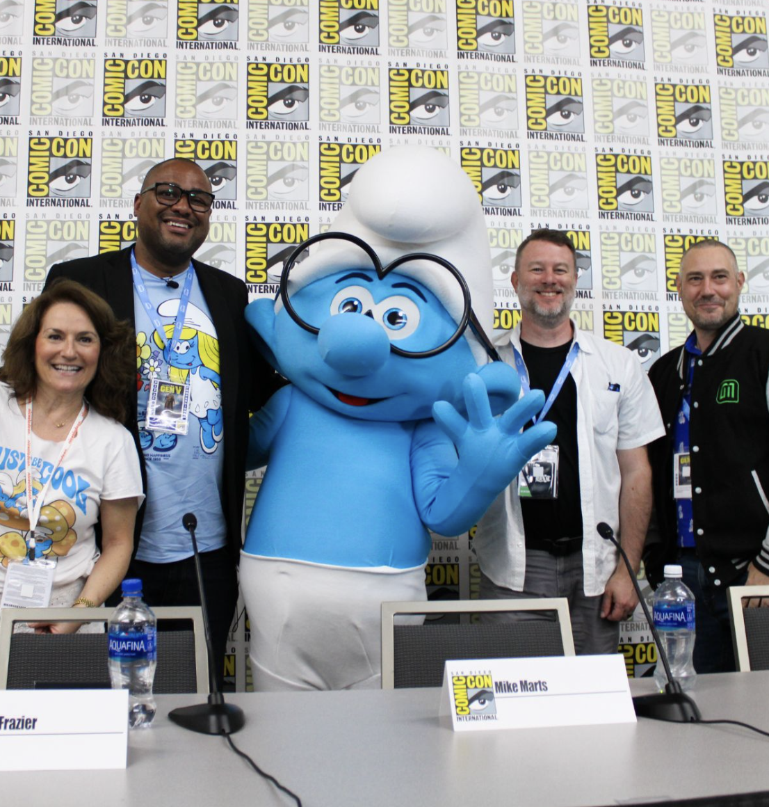 Group photo at Comic-Con featuring a large Smurf mascot with glasses flanked by four people, two men and two women, standing behind a conference table with name tags and water bottles, backdrop with Comic-Con logos.