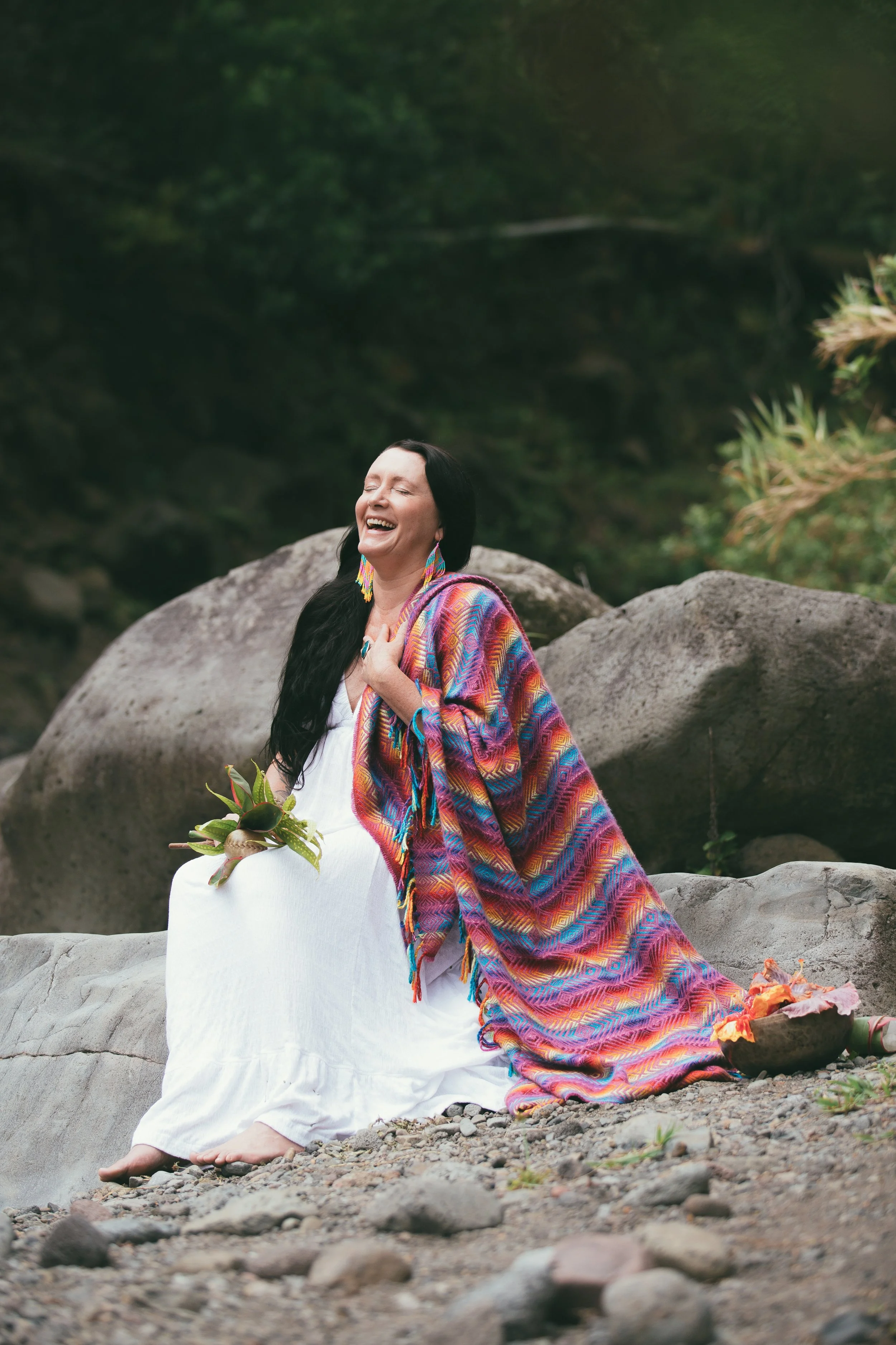 A joyful woman with long dark hair, sitting on rocks outdoors, wearing a white dress, a colorful shawl, and earrings, holding a small plant, with a background of trees and greenery.
