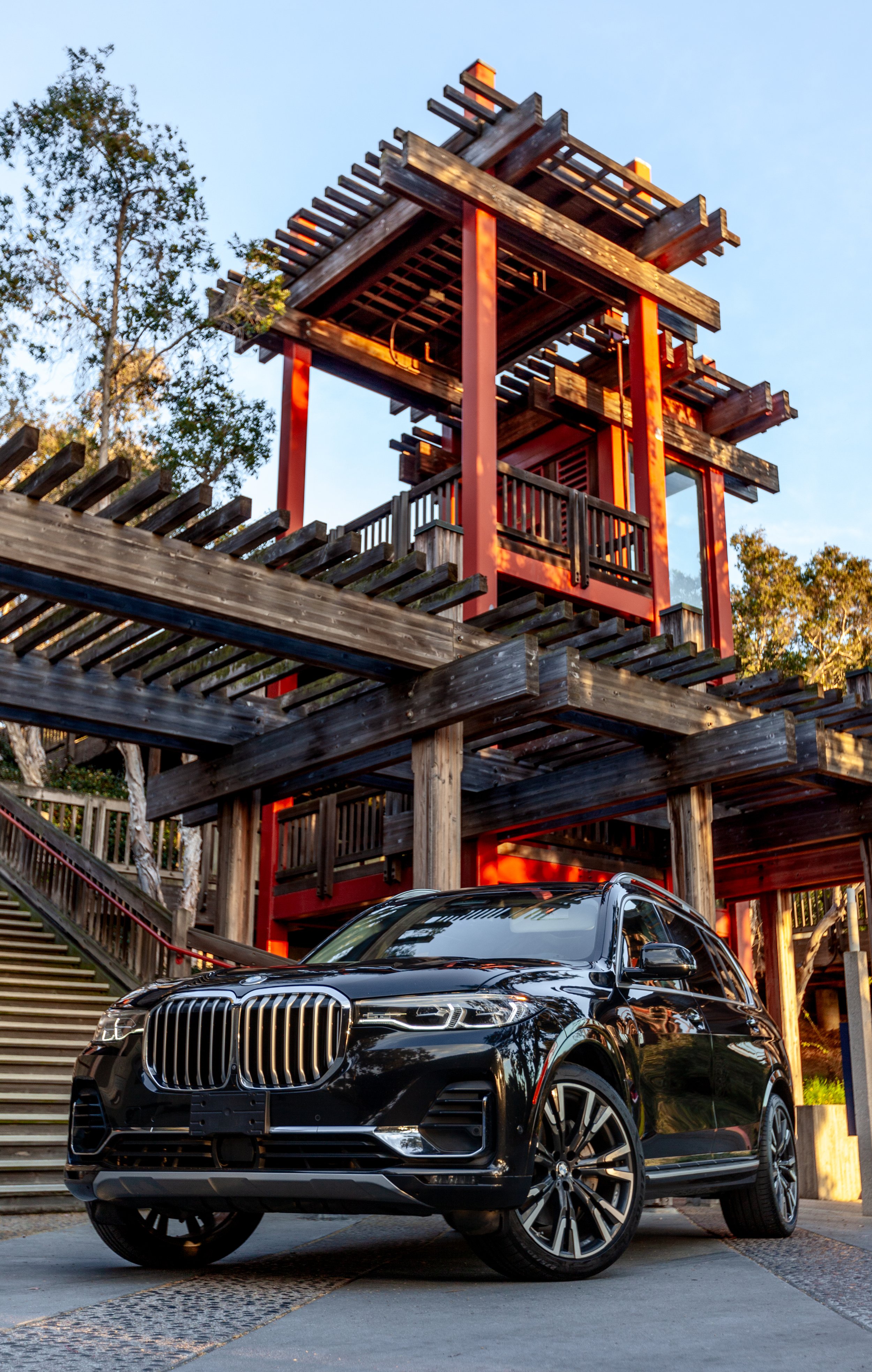 A black luxury SUV parked in front of a multi-level wooden treehouse with red support beams during sunset.