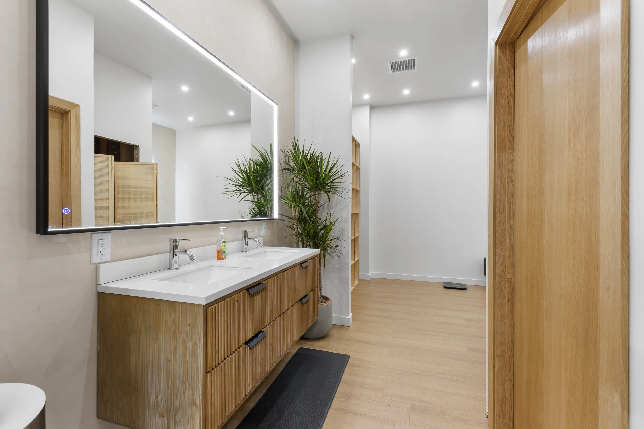 Bathroom with dual sinks, wooden vanity, large illuminated mirror, potted plant, and wooden sliding door, with wood flooring and recessed ceiling lights.