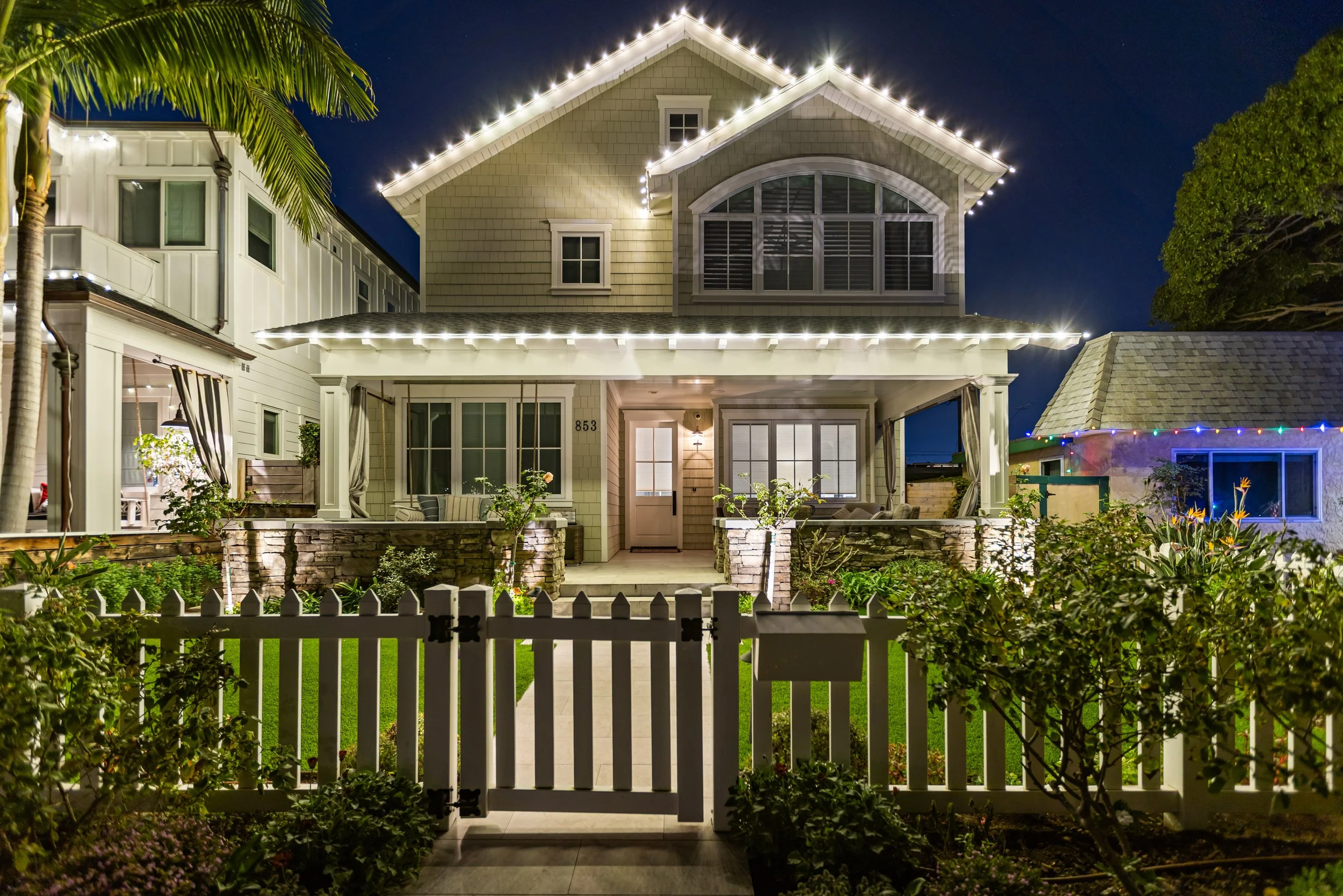 Night view of a house decorated with Christmas lights, with a white picket fence and garden in the front yard.