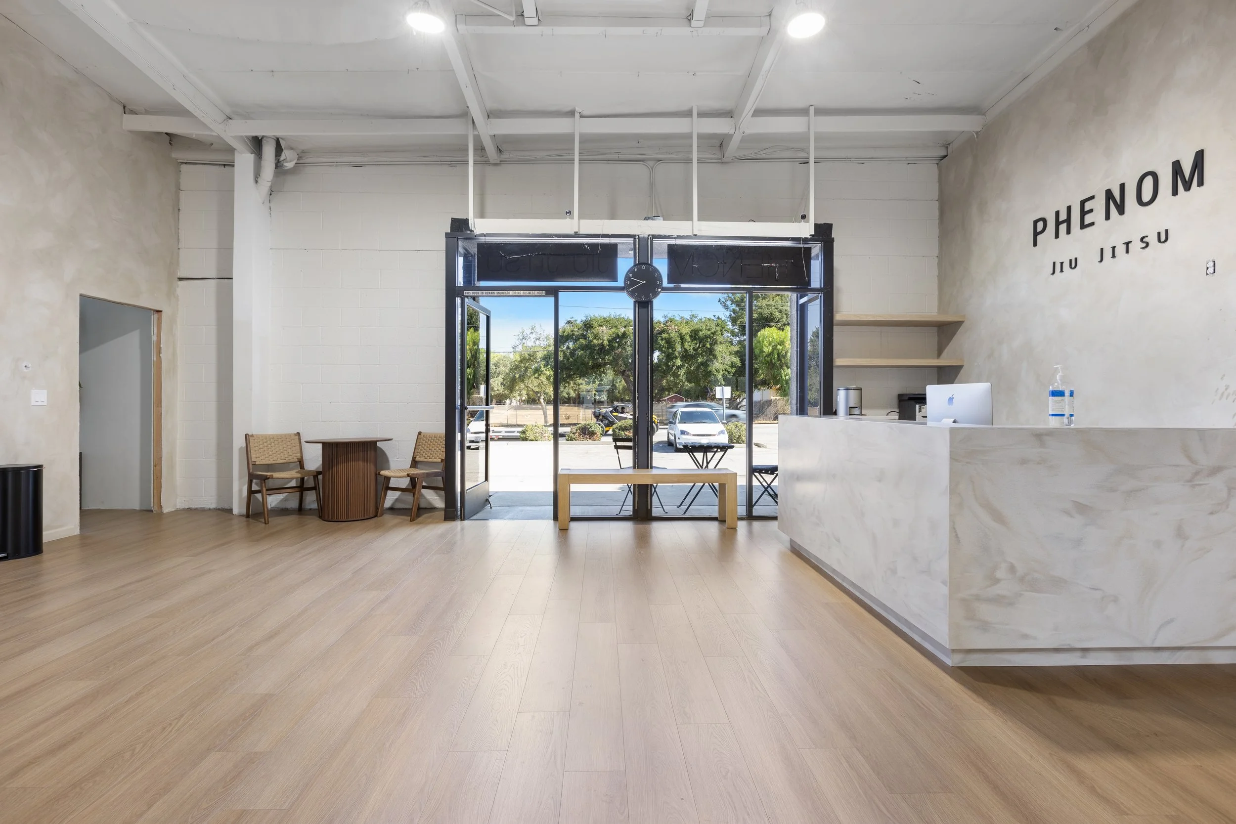 Empty martial arts studio with a reception desk, chairs, and a glass front door showing outdoor trees and parking lot.