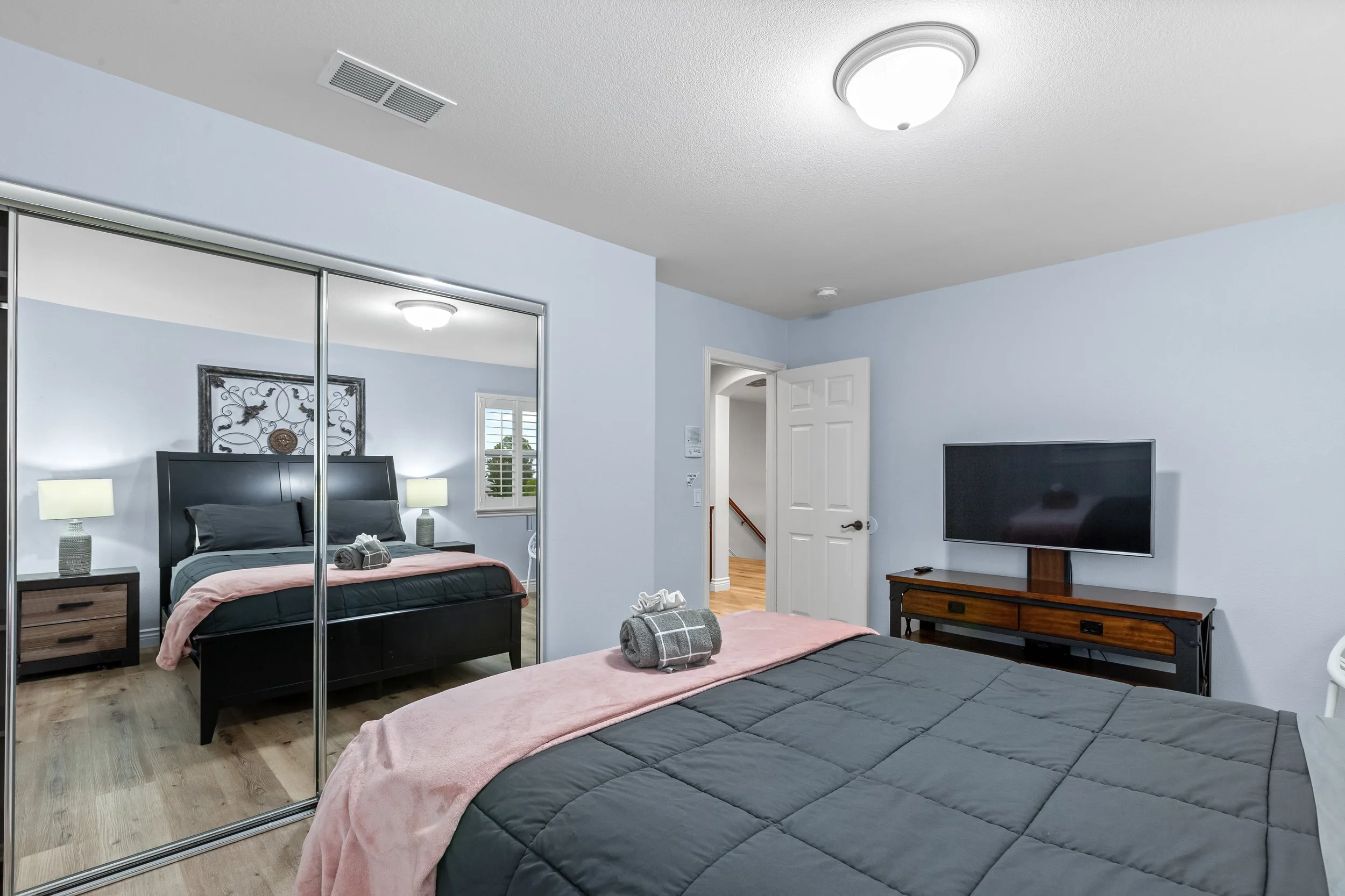 A bedroom with a bed, bedside tables with lamps, a closet with mirrored sliding doors, a television on a wooden stand, a door to an adjacent room, and light blue walls.
