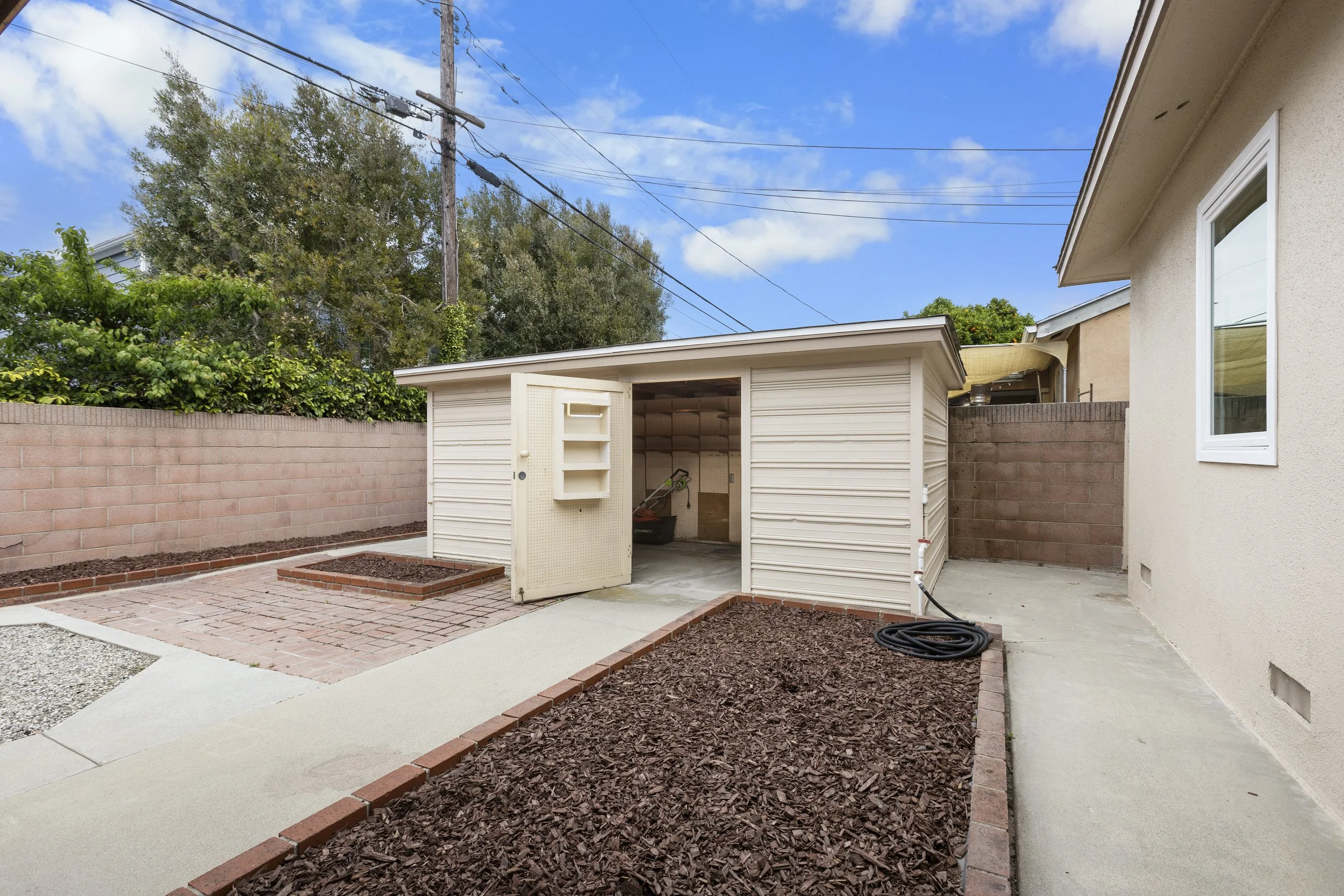 Backyard with a beige shed, brick and concrete patio, and newly mulched garden beds, surrounded by a brick wall and a house with beige stucco exterior.