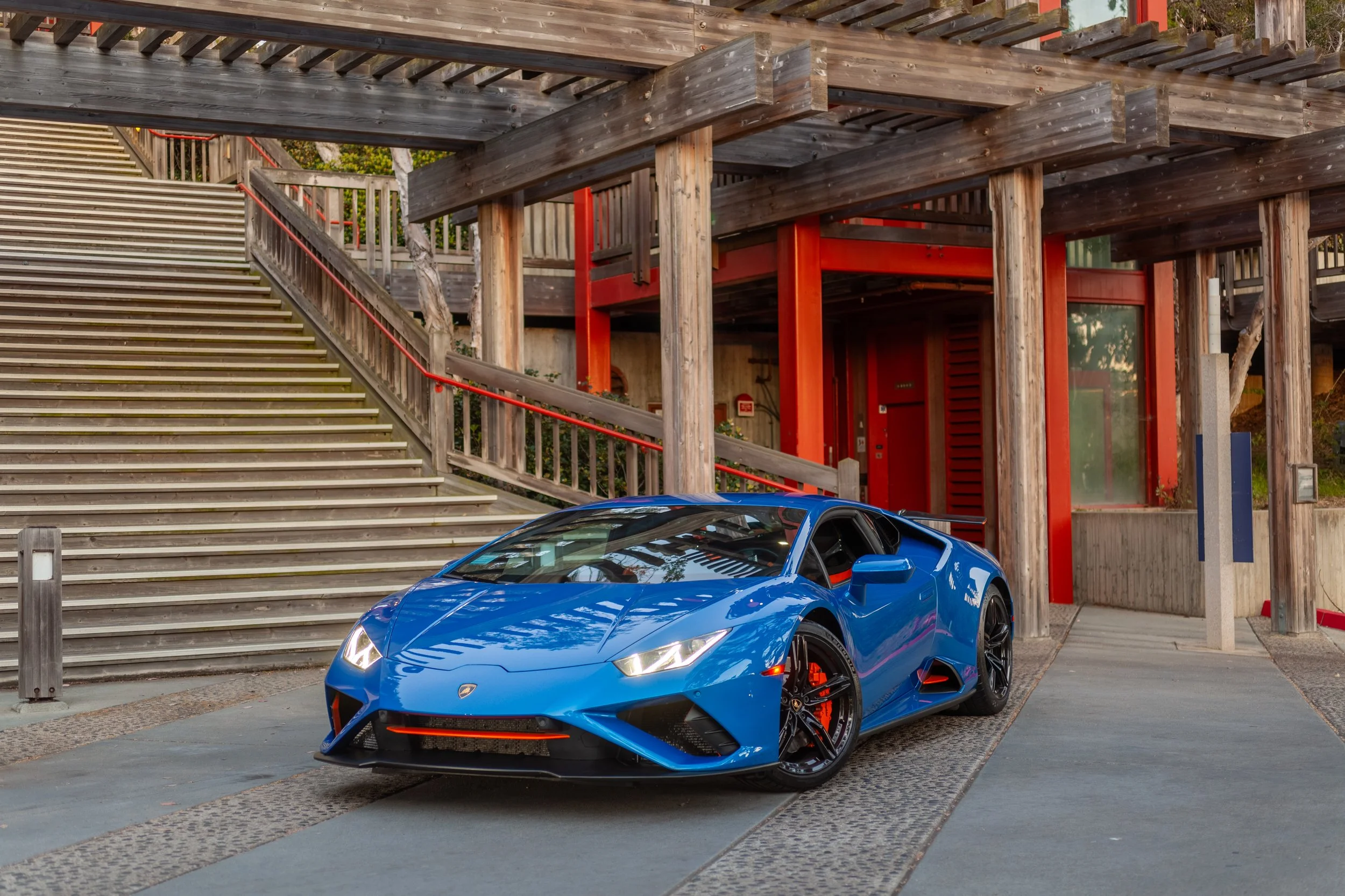 A blue Lamborghini sports car parked on a paved area with a multi-level wooden stairway and building in the background.