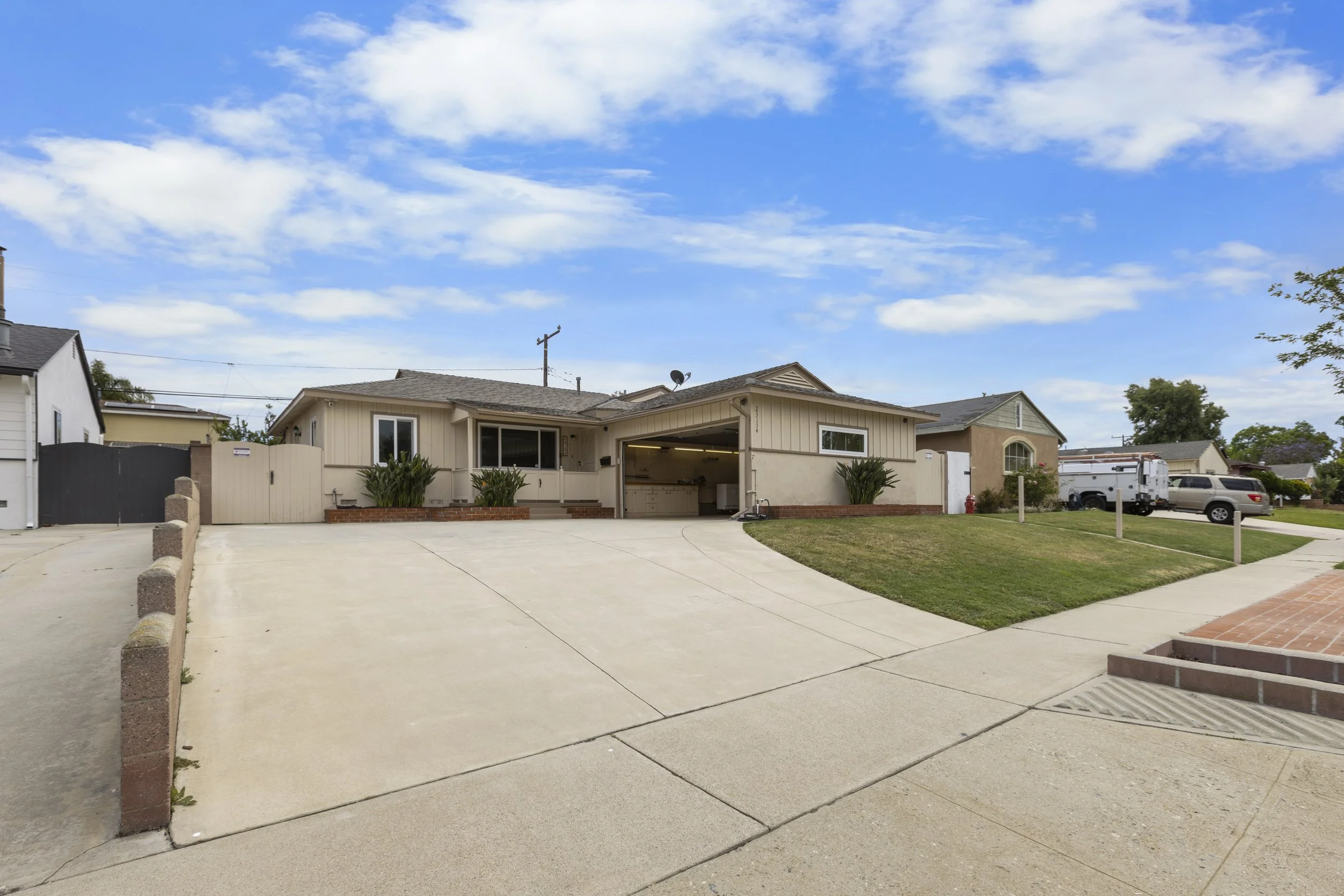 A beige house with an attached garage on a residential street, with a large concrete driveway and a grassy lawn, under a partly cloudy blue sky.