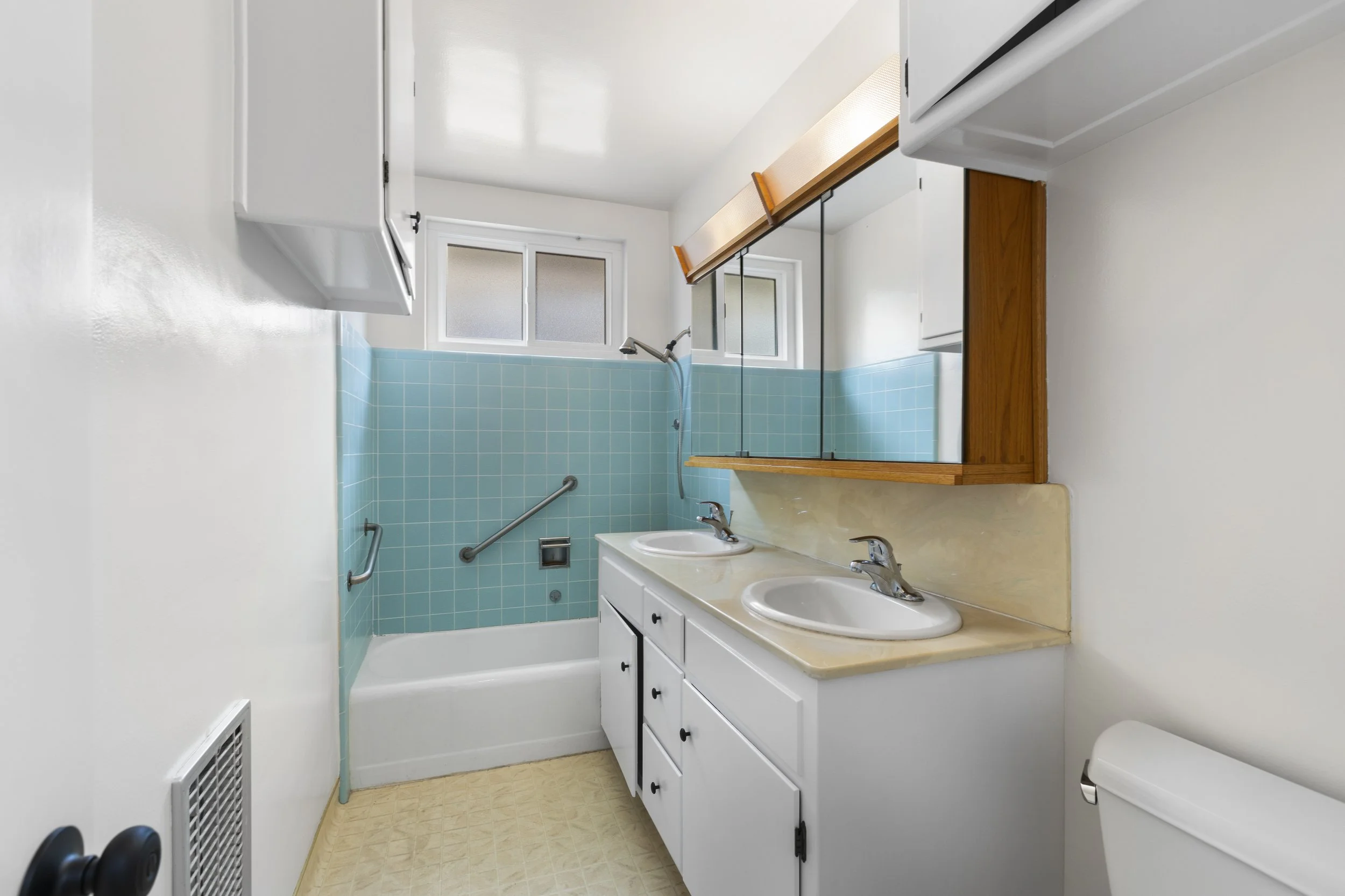 Small bathroom with double vanity sinks, mirror cabinets, a blue-tiled bathtub, and a window with frosted glass.