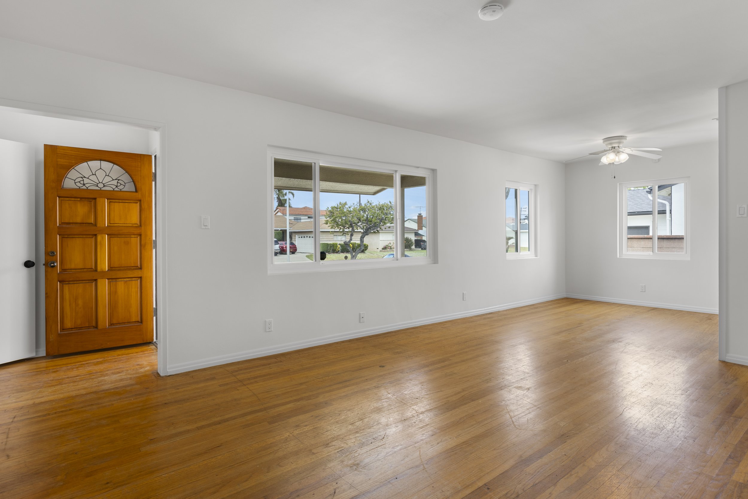 Empty living room with white walls, hardwood floors, multiple windows, a ceiling fan, and a wooden front door.