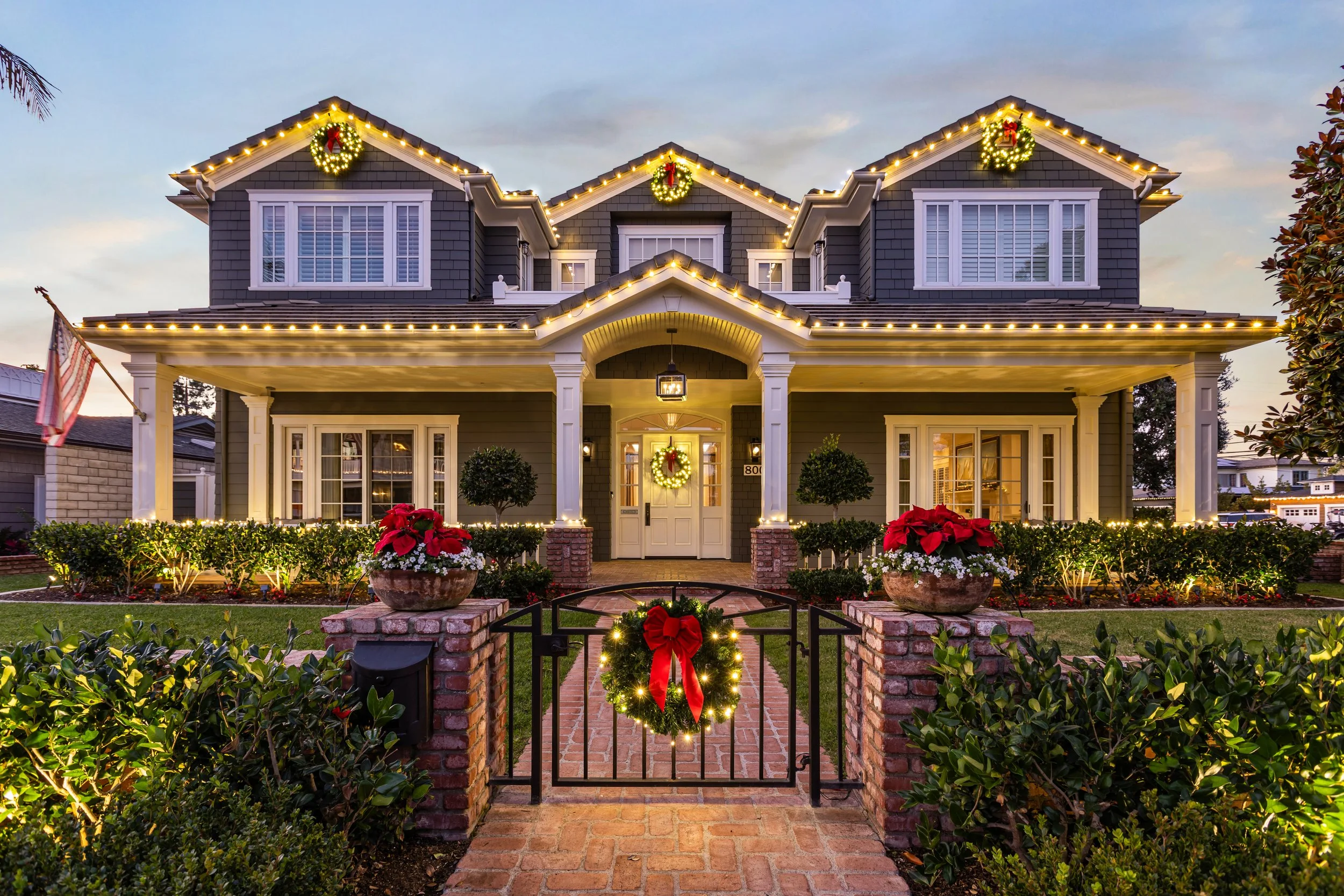 A house decorated for Christmas with lights, wreaths, poinsettias, and festive wreath on the front door, with a brick pathway and a black gate decorated with a Christmas wreath and red bow.