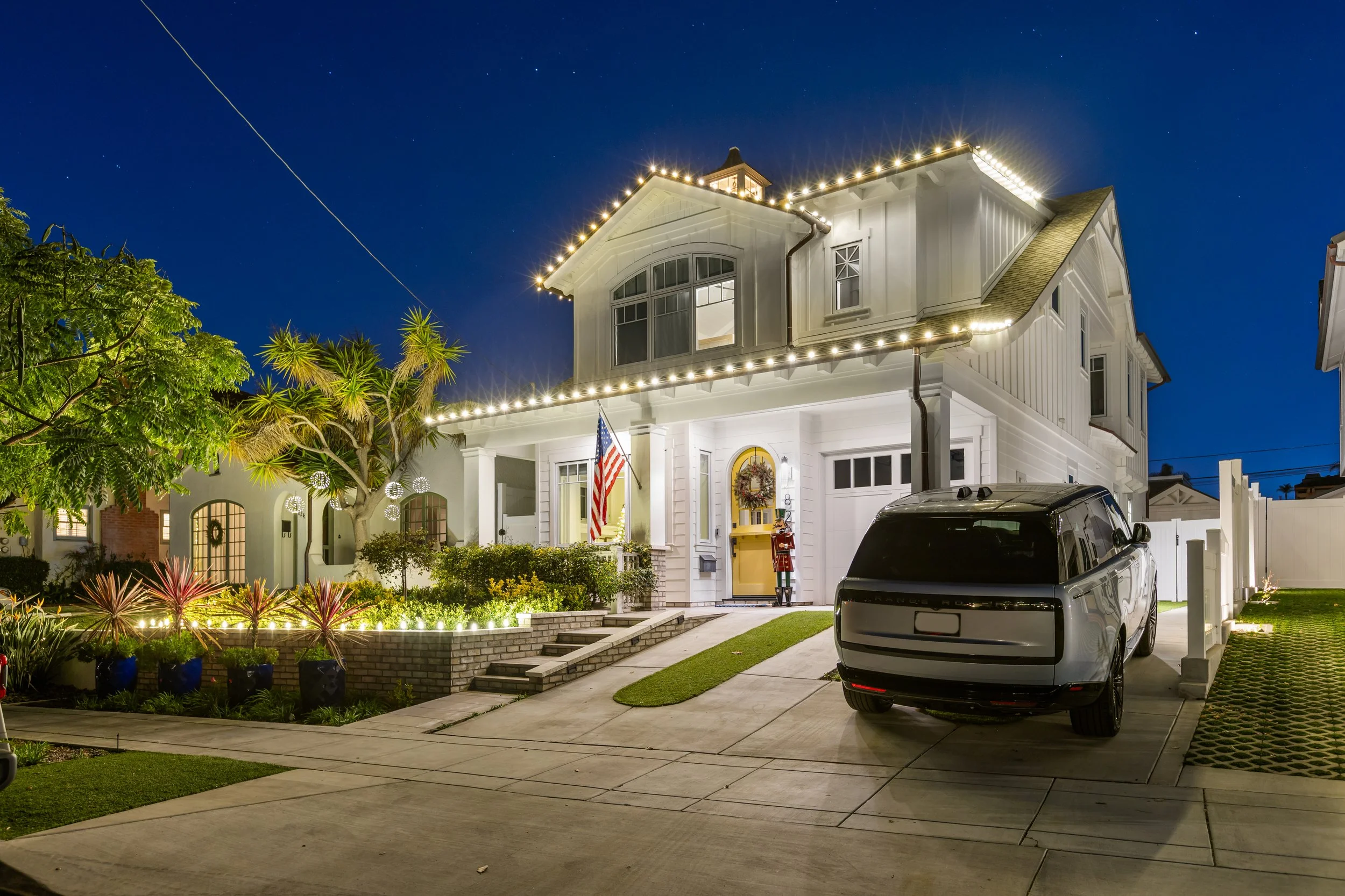 A two-story white house decorated with string Christmas lights along the roofline, with a front porch, American flag, and ornaments. A gray and black SUV is parked in the driveway at night, with planting beds, trees, and a night sky in the background.