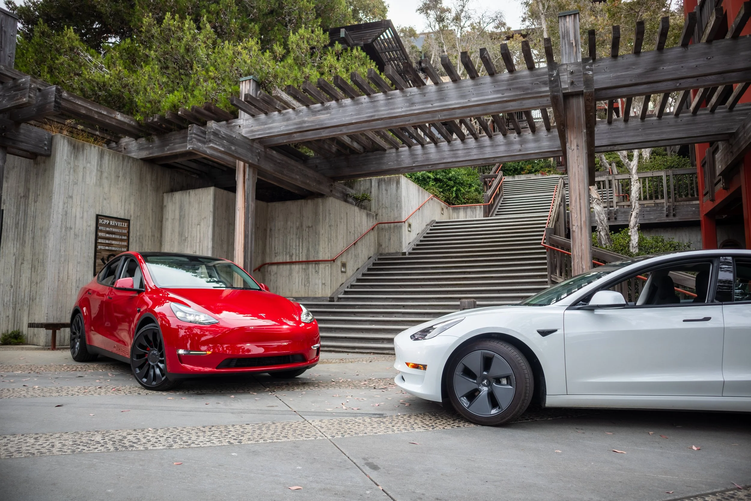 A red Tesla and a white Tesla parked under a wooden structure near stairs in an outdoor setting.