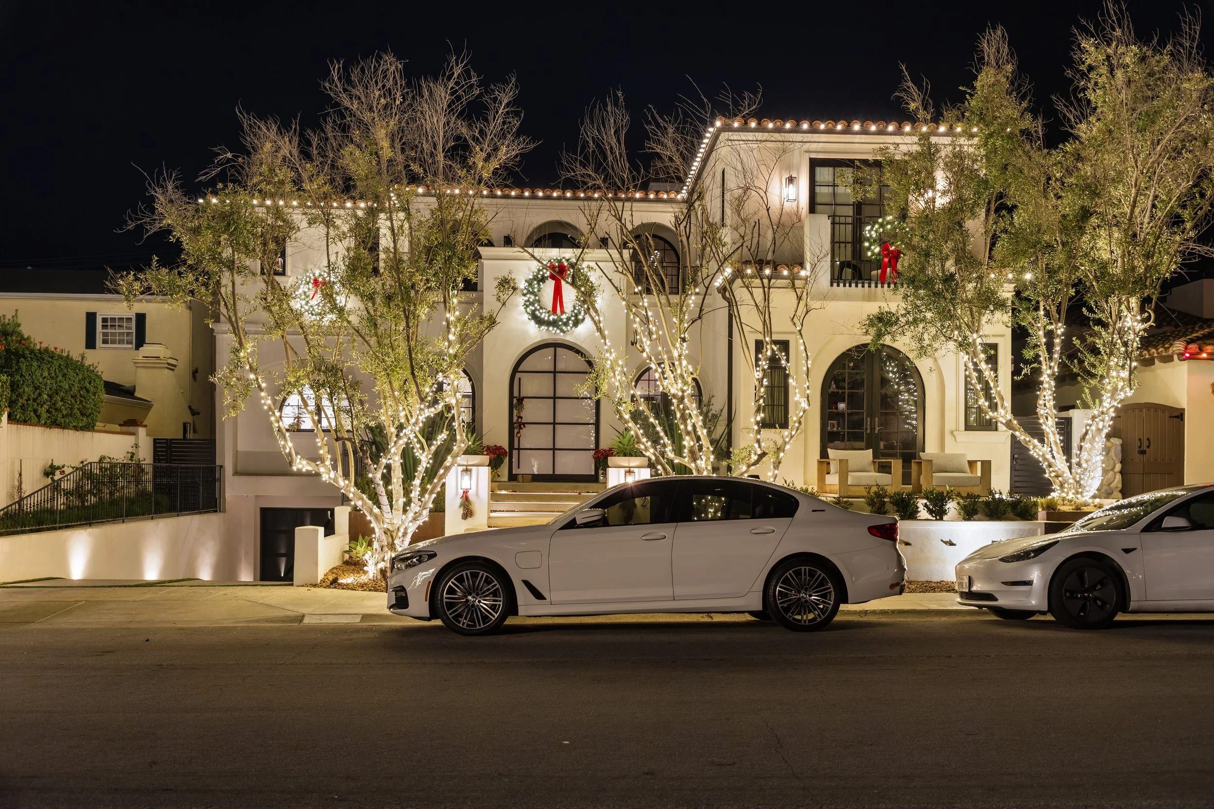 A house decorated with Christmas lights and wreaths at night, with two parked cars in front.