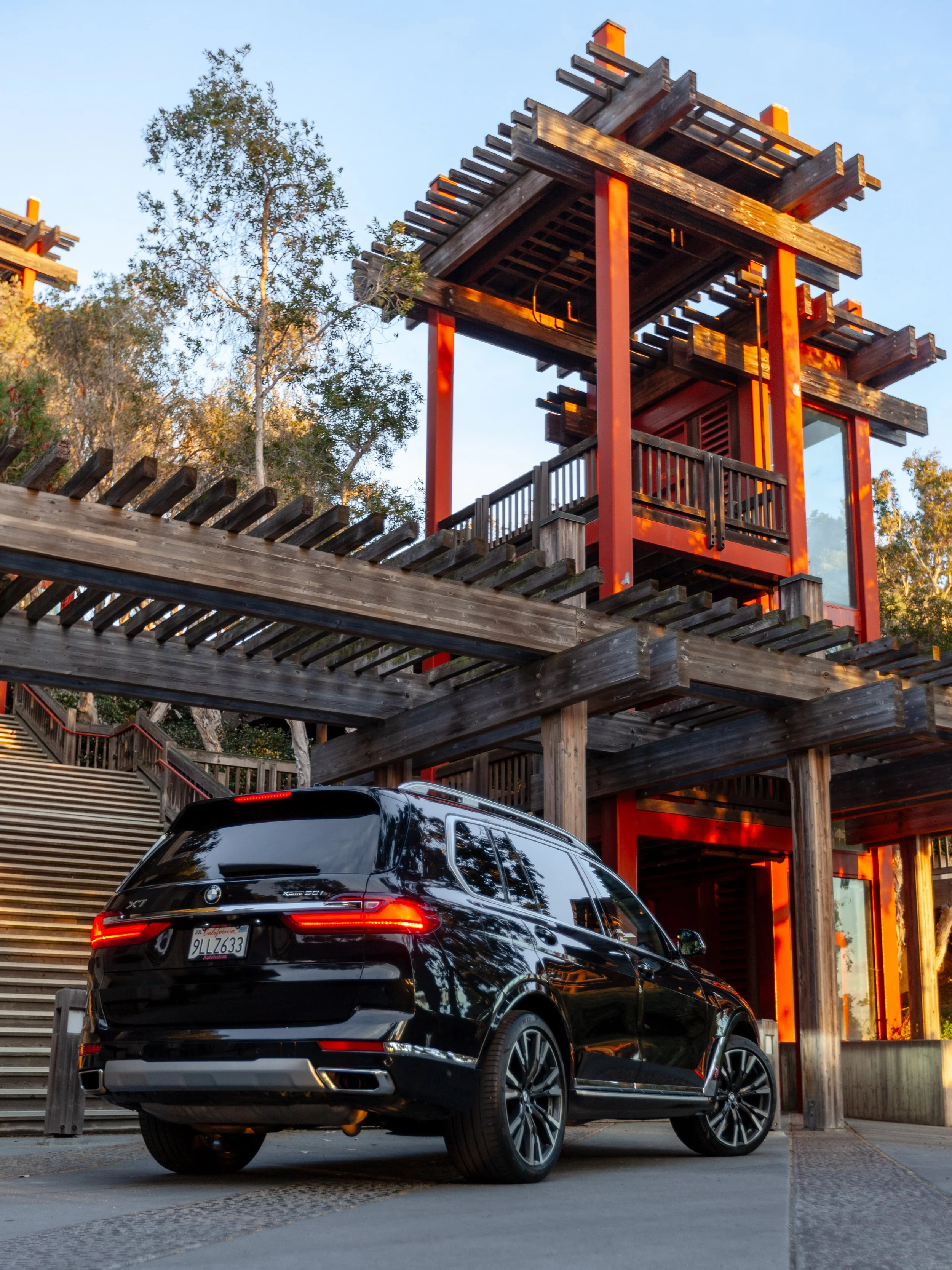 A black luxury car parked in front of a modern multi-level wooden structure with red accents, surrounded by trees and clear blue sky.