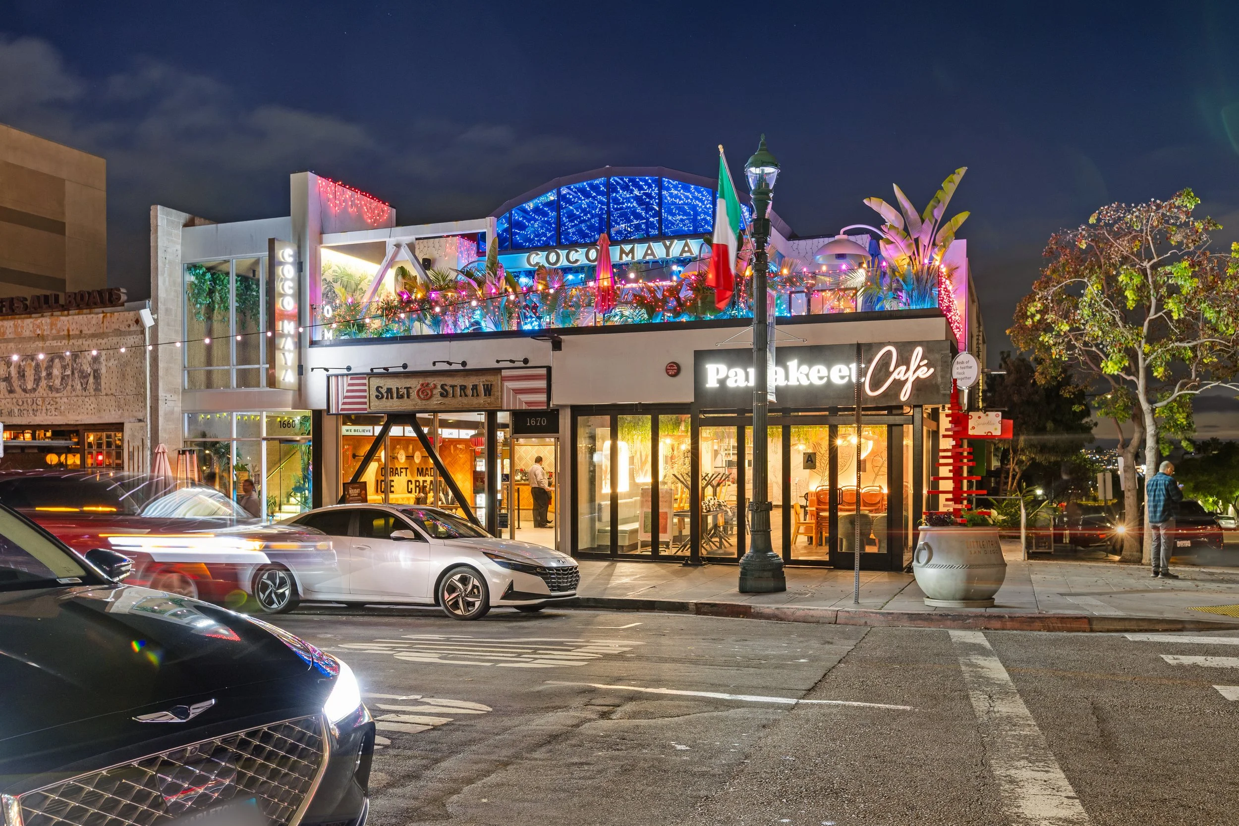 Nighttime street scene featuring a two-story building with a restaurant on the ground floor named 'Pakakee Café' and a decorated rooftop area with colorful lights, plants, and a sign that reads 'Coco Maya'. Cars are parked in front and some are moving, creating light trails. A man is walking on the sidewalk next to a tree.