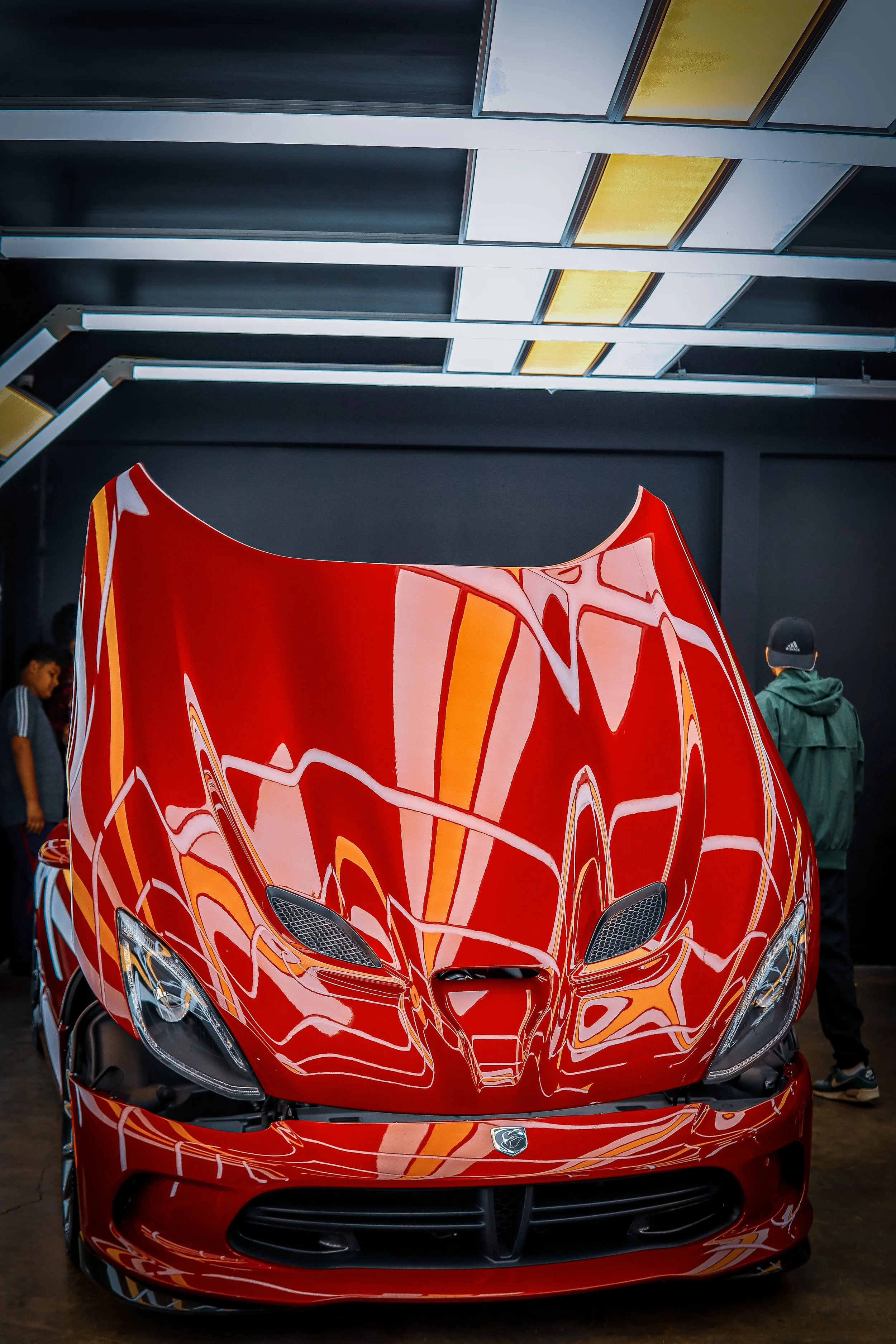 Red sports car on display in a showroom with reflective ceiling panels and people observing it.