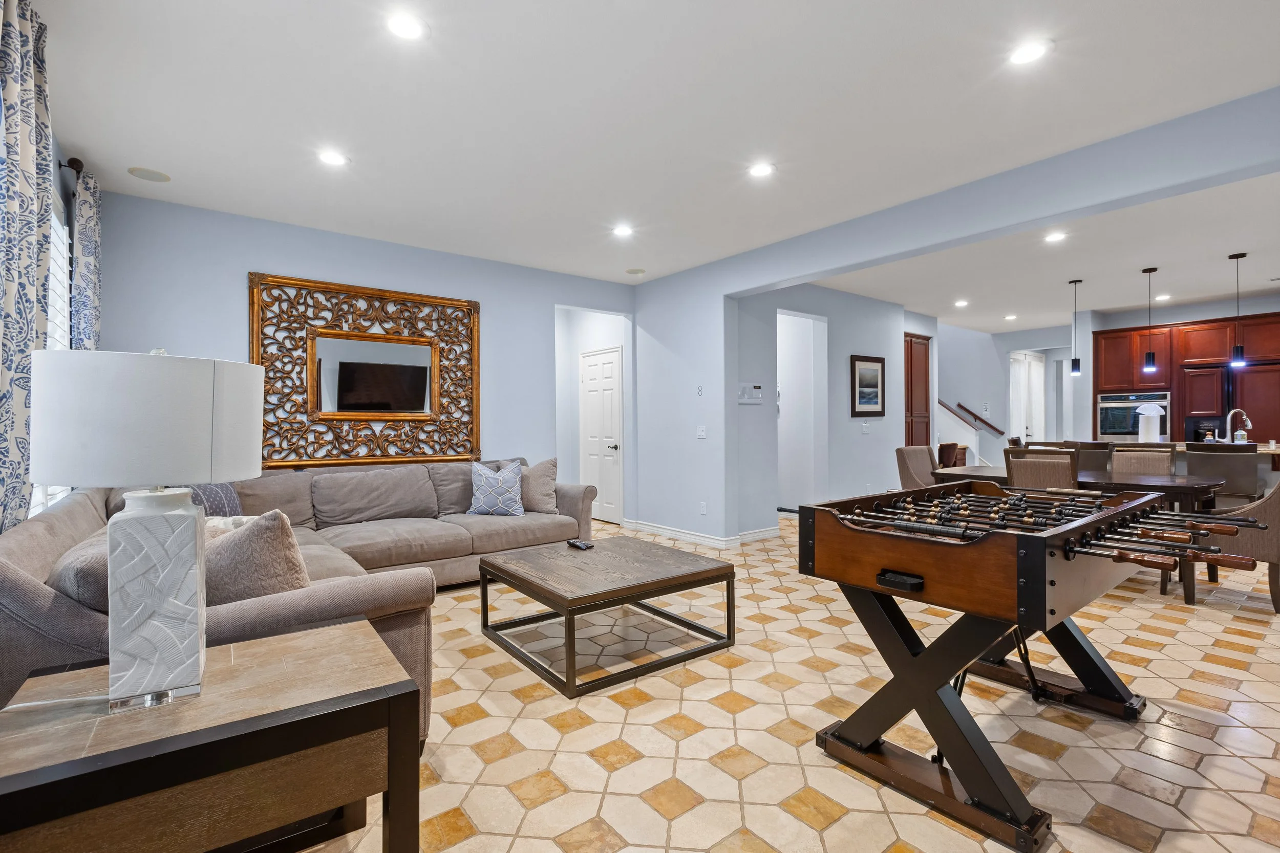 Living room with beige sofas, a wooden coffee table, a foosball table, and a dining area in the background with a hanging light fixture, dark wood cabinets, and kitchen appliances.