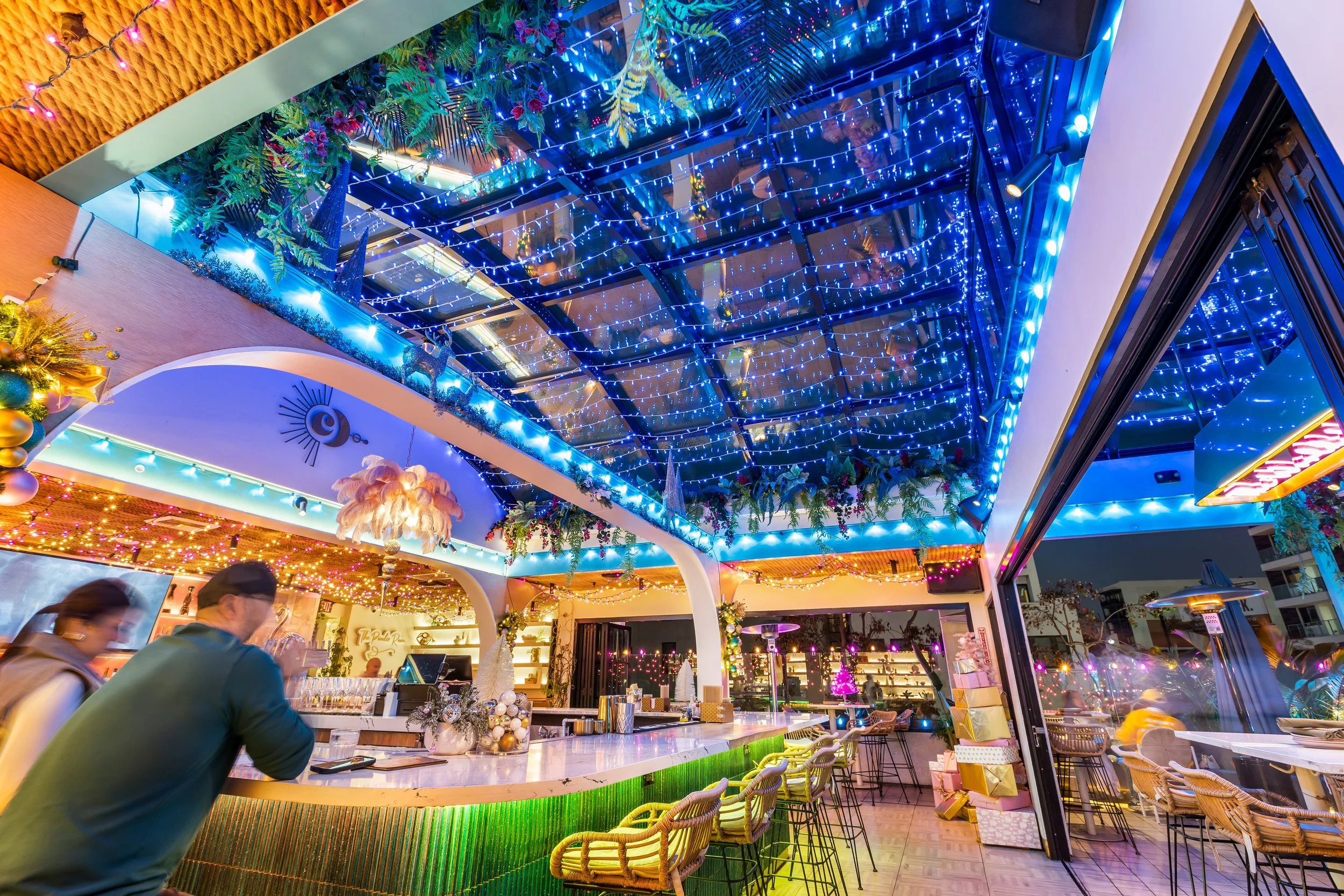 Interior of a festive bar or restaurant decorated with colorful holiday lights, ornaments, and greenery, featuring a curved bar counter, seating, and a glass ceiling with blue string lights.