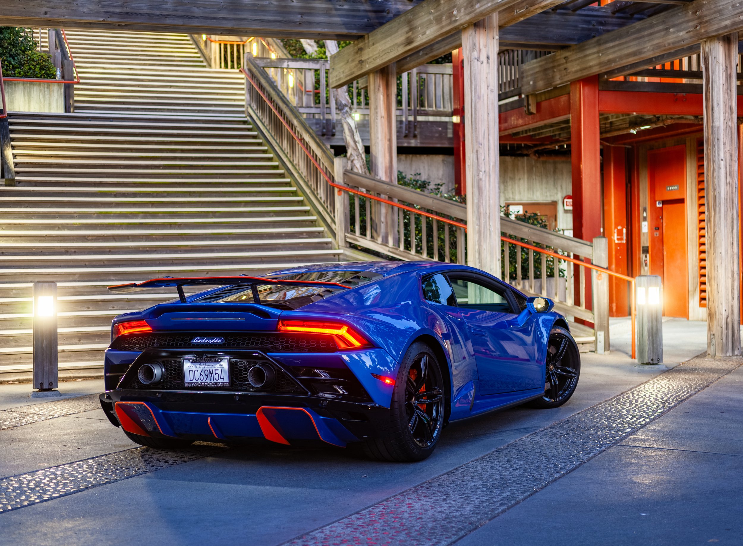 A blue Lamborghini sports car parked near a wooden staircase with railings, illuminated by nearby lights at dusk or evening.