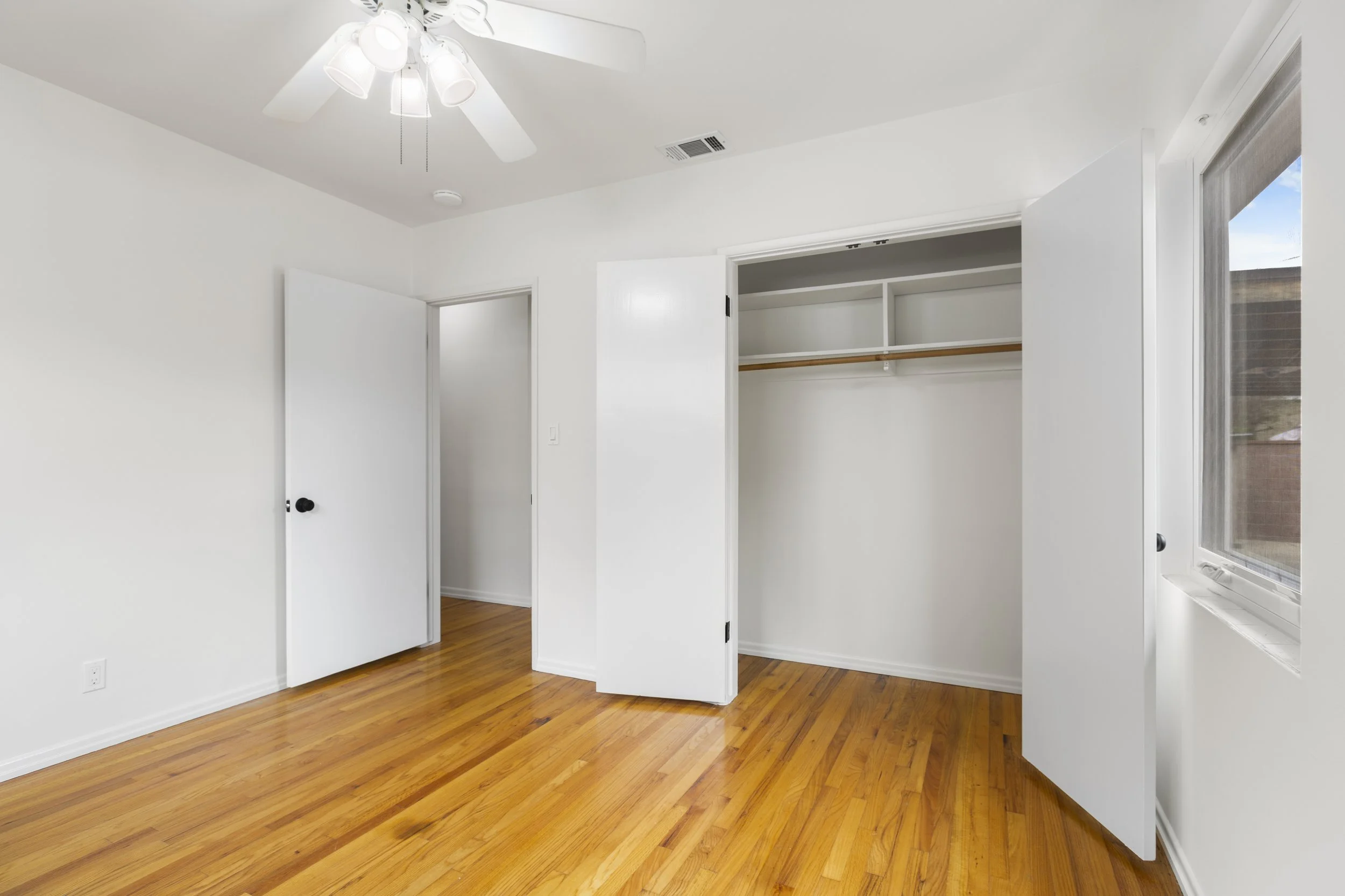 Empty room with hardwood floors, white walls, a ceiling fan, a closet with open doors, and a window.