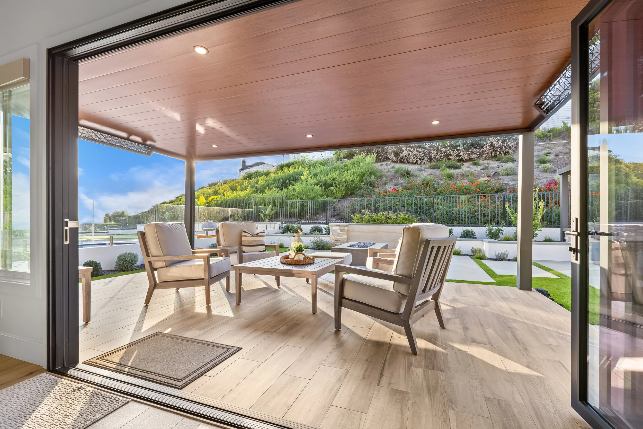 View of an outdoor patio with beige chairs and a coffee table, overlooking a landscaped yard with greenery, flowers, and a hillside with trees under a clear blue sky.