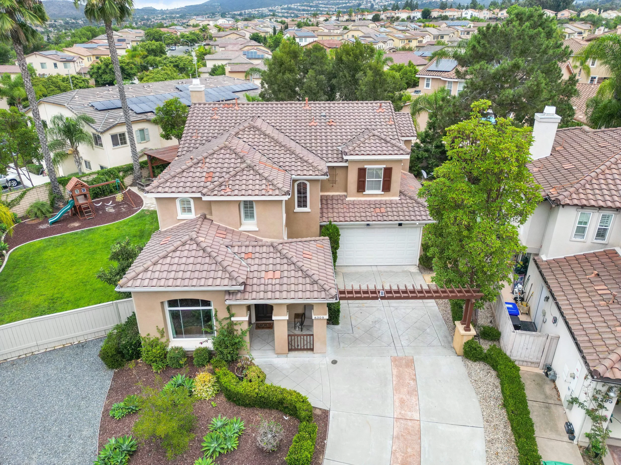 Aerial view of a suburban house with a tiled roof, front yard with bushes and trees, driveway, and neighboring houses with similar roofs, green lawns, and trees. There's a children's play area with a slide in the backyard.