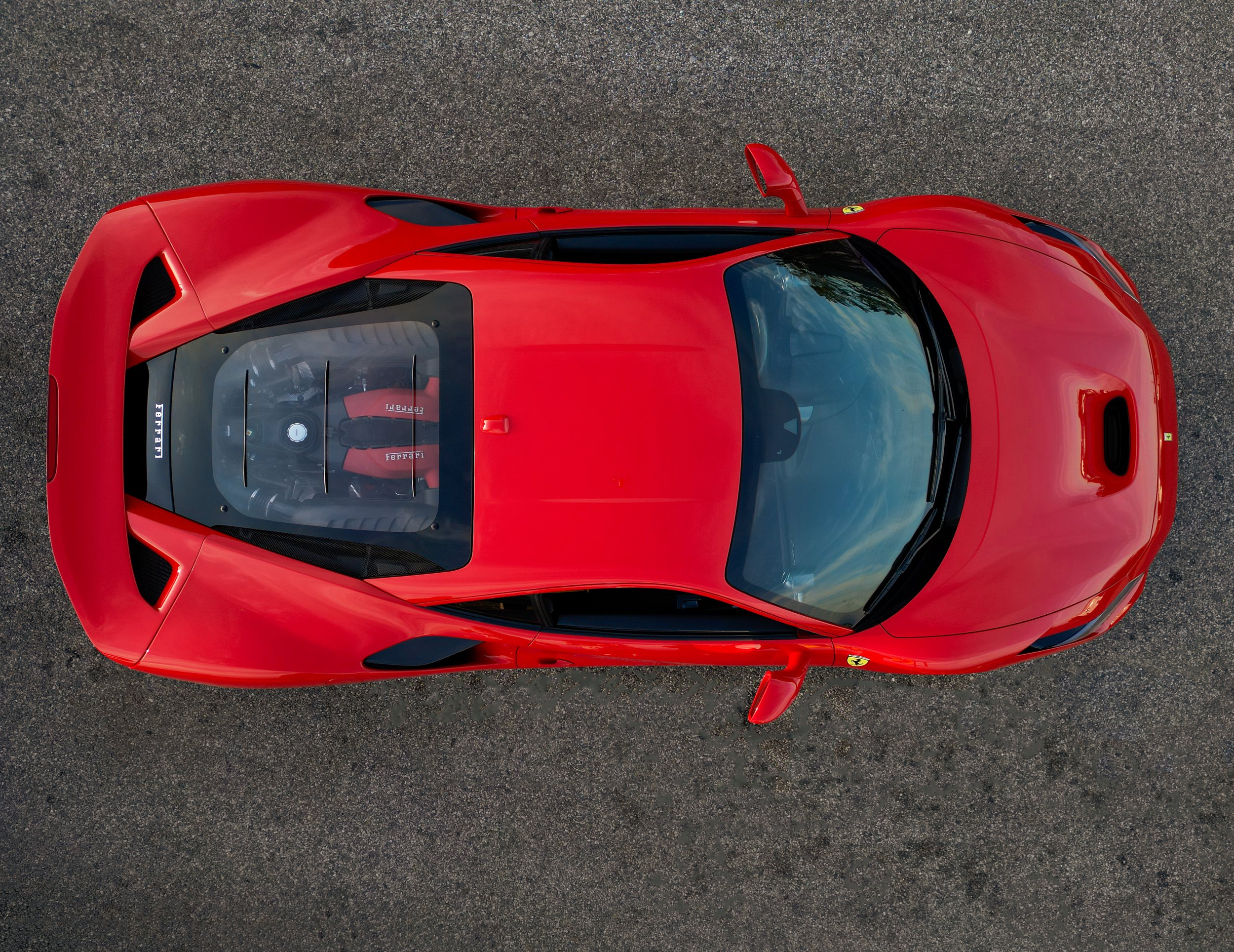 Top view of a red Ferrari sports car parked on asphalt, showing the engine through a transparent cover on the rear.