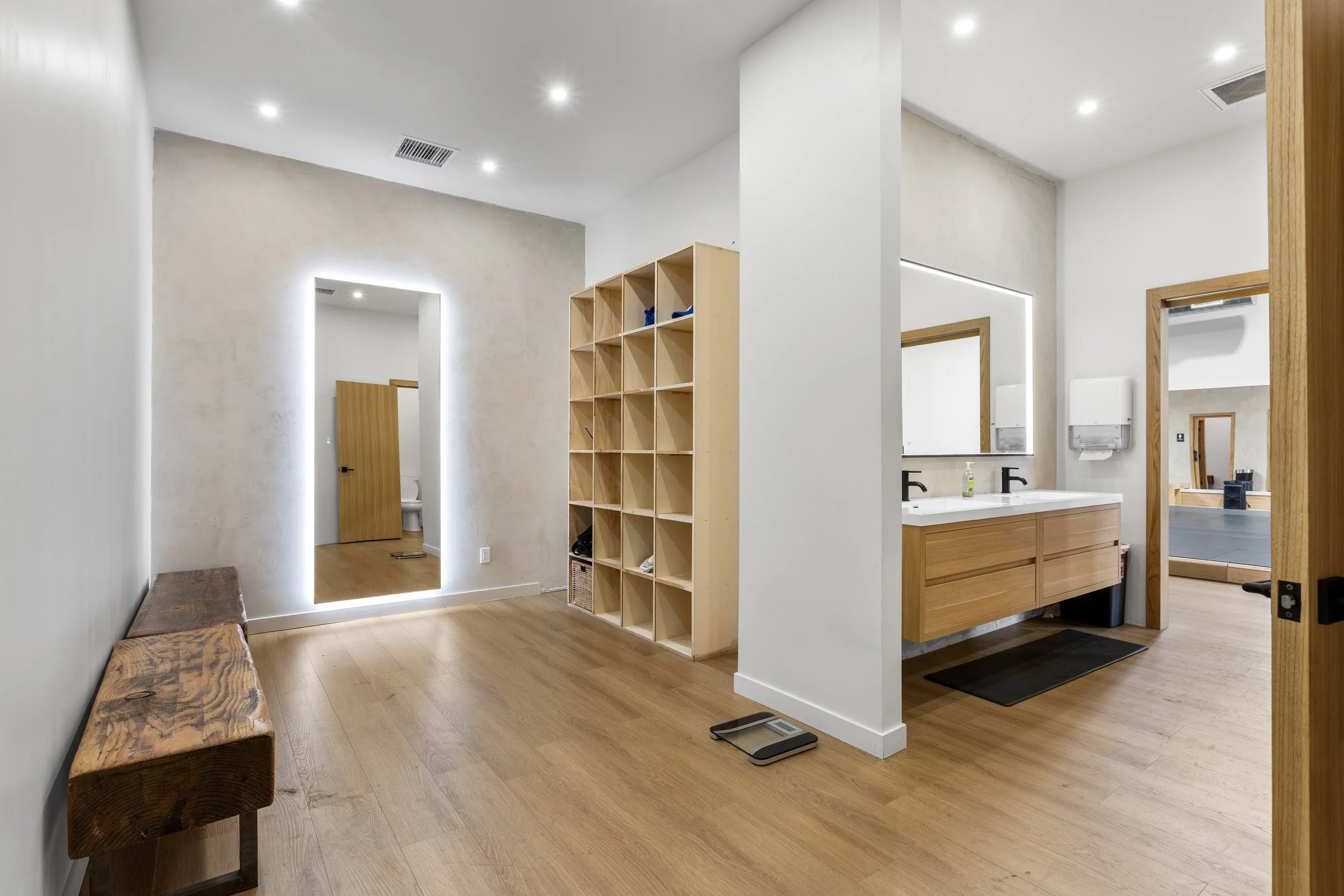 Interior of a modern, minimalist room with wooden floors, a wooden bench, a large mirror with LED lighting, a wooden cubby shelving unit, a bathroom sink with a large mirror, and a small adjoining room with a ping pong table.