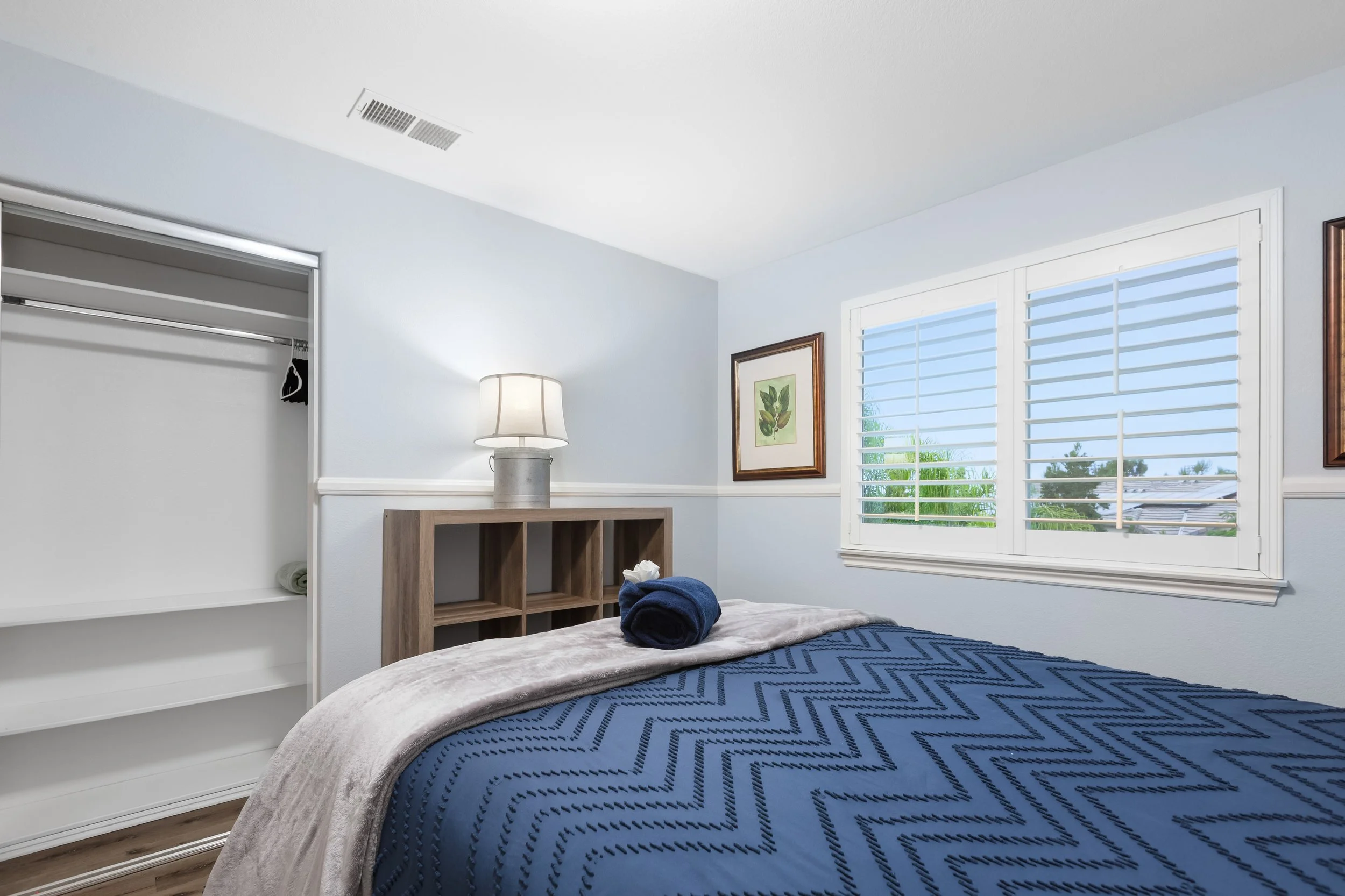 A bedroom with a bed covered in a dark blue quilt, a blanket and towel rolled on top. There's a window with white shutters and a framed botanical print on the wall. An open closet with some hanging clothes is visible. A wooden nightstand with cubbies and a lamp is next to the bed.