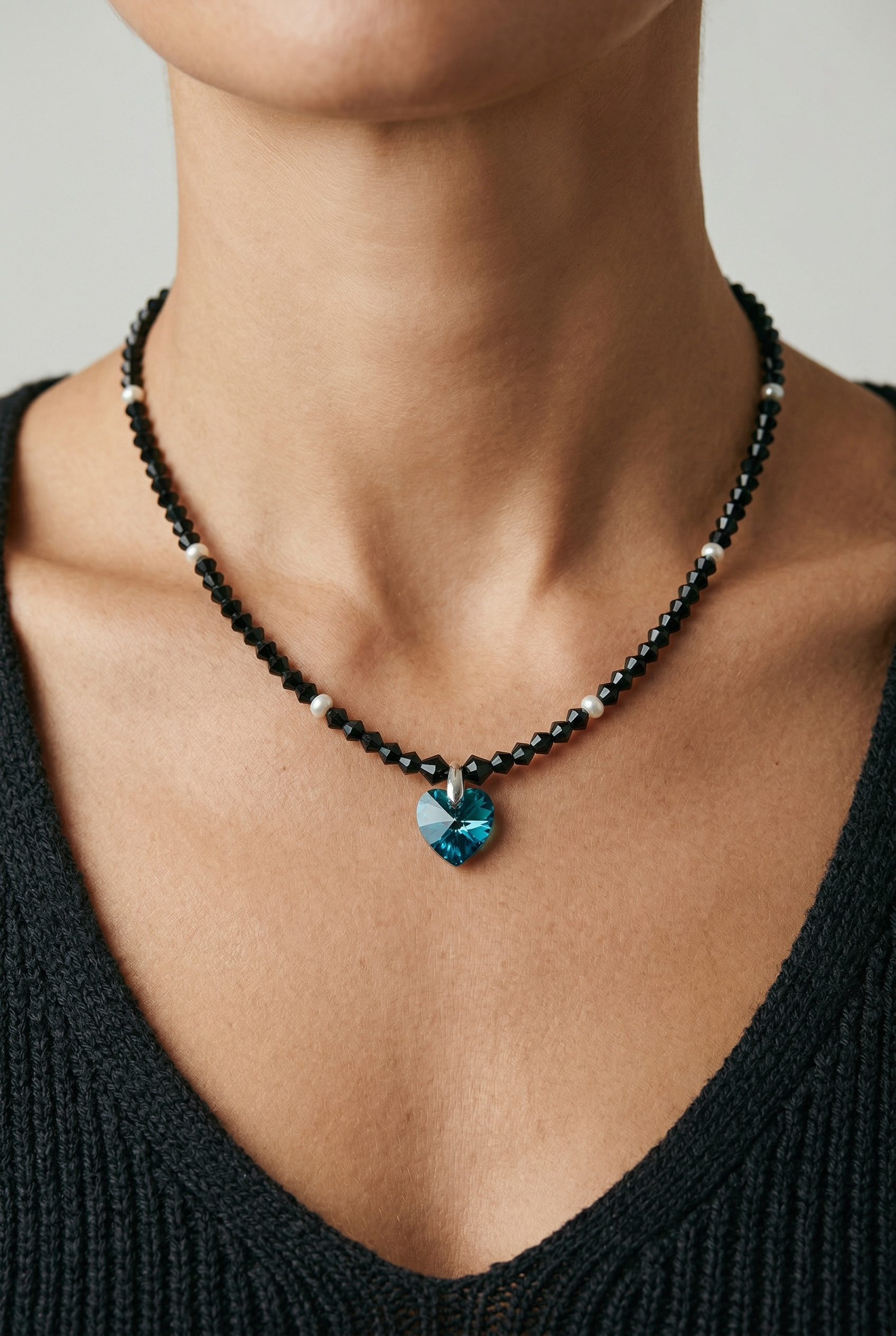 Close-up of a woman wearing a black beaded necklace with small pearl accents and a large heart-shaped blue gemstone pendant.