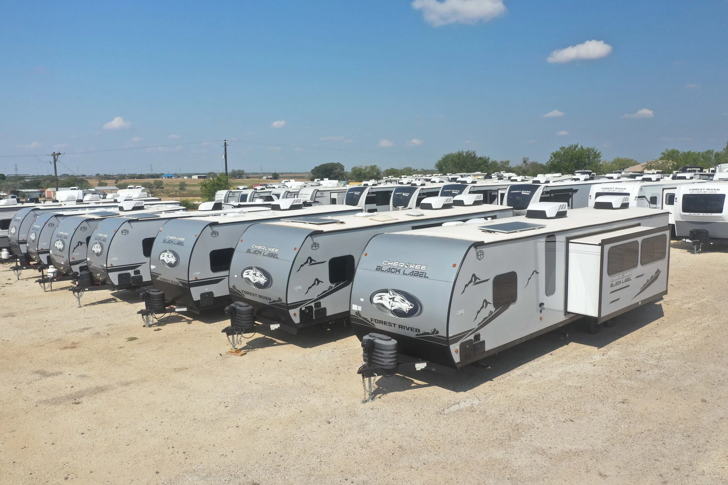 A large lot filled with rows of identical white travel trailers with mountain and animal graphics, under a blue sky with scattered clouds.