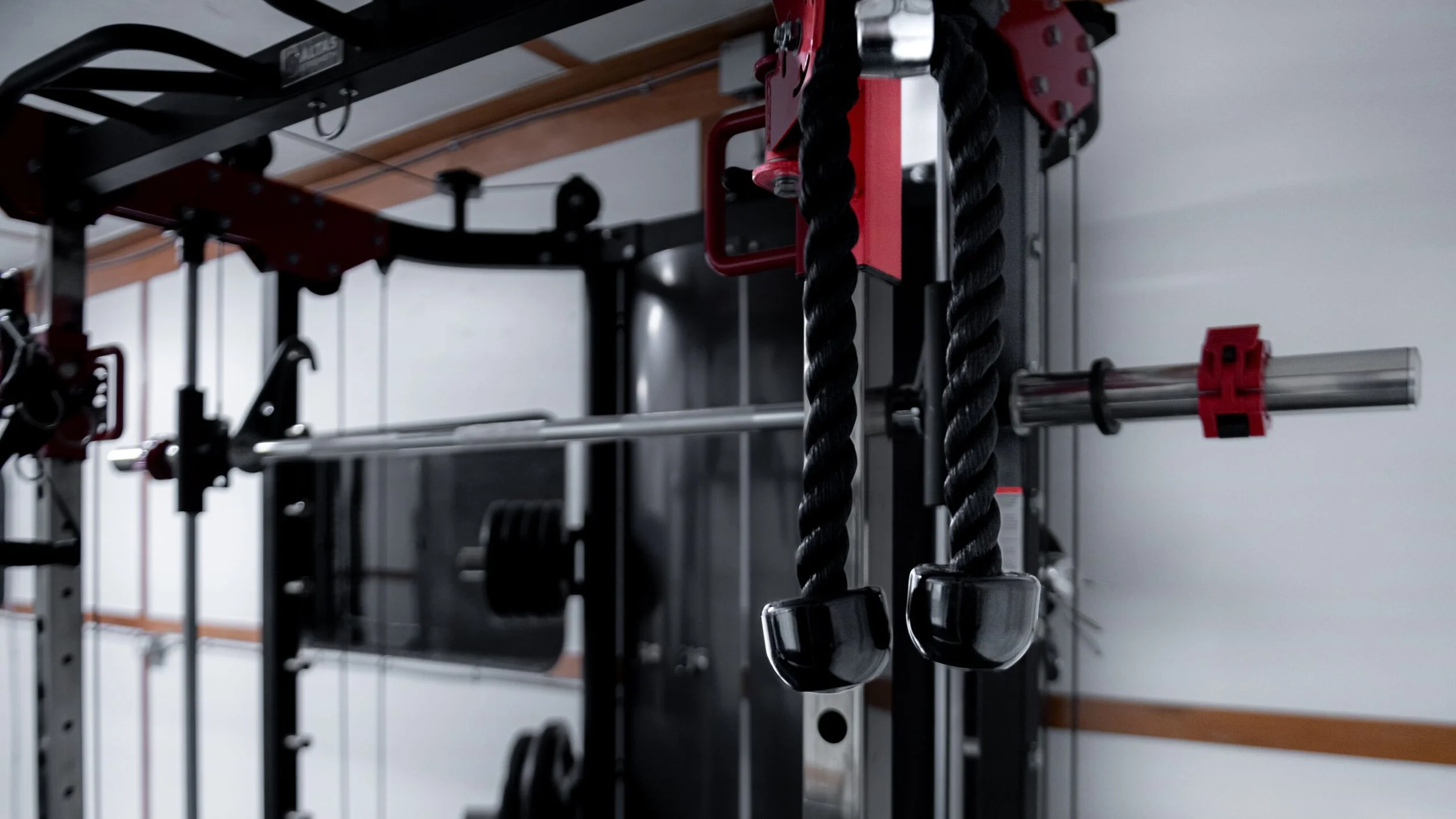 Close-up of a gym or fitness equipment setup, featuring black battle ropes with plastic caps, a metal barbell, and a pulley system with red accents in a well-lit room with a white wall background.