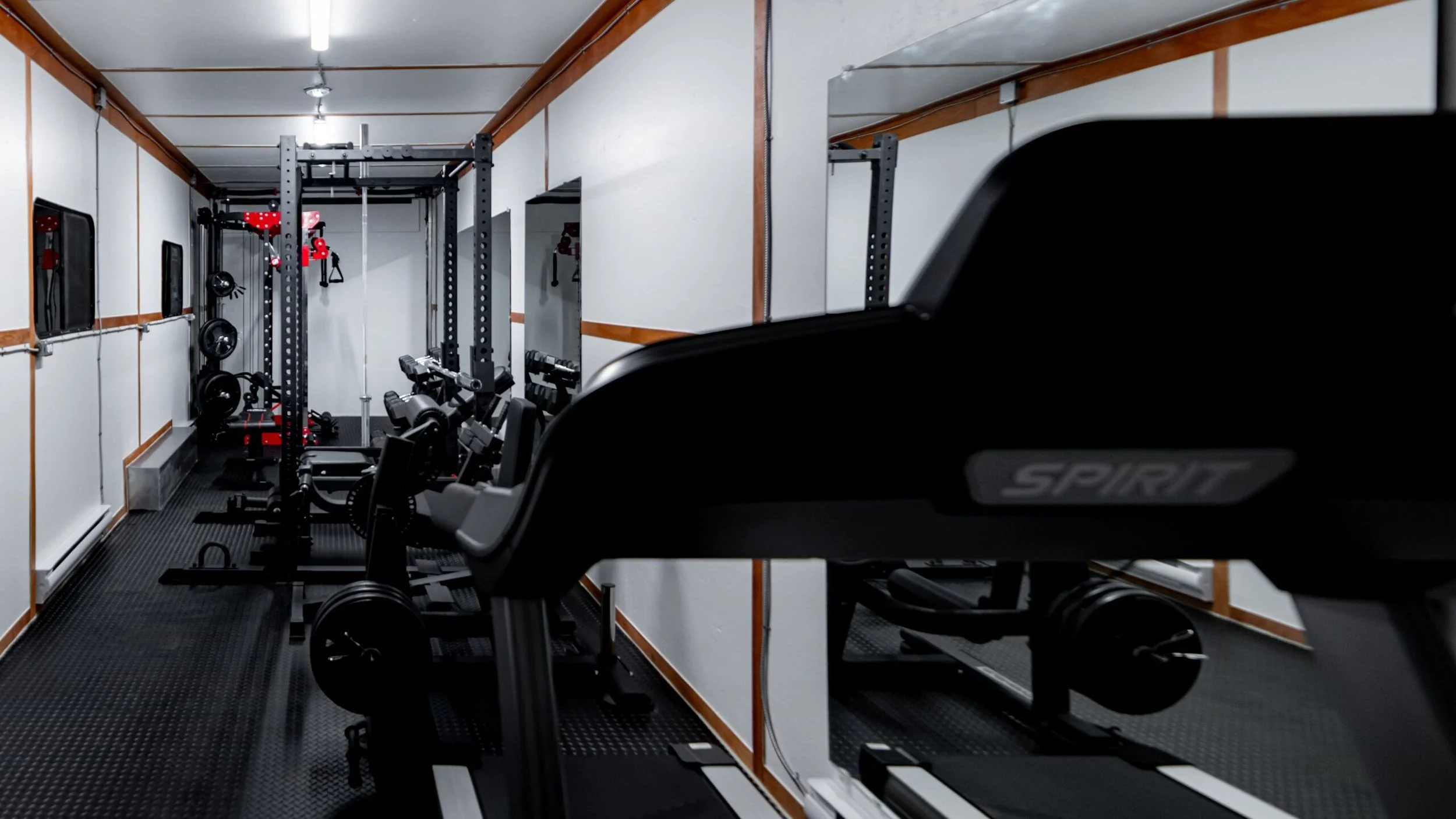 A small, well-lit gym with various workout equipment including dumbbells, weight racks, and exercise machines. The room has white walls with wood trim and a black rubber floor.