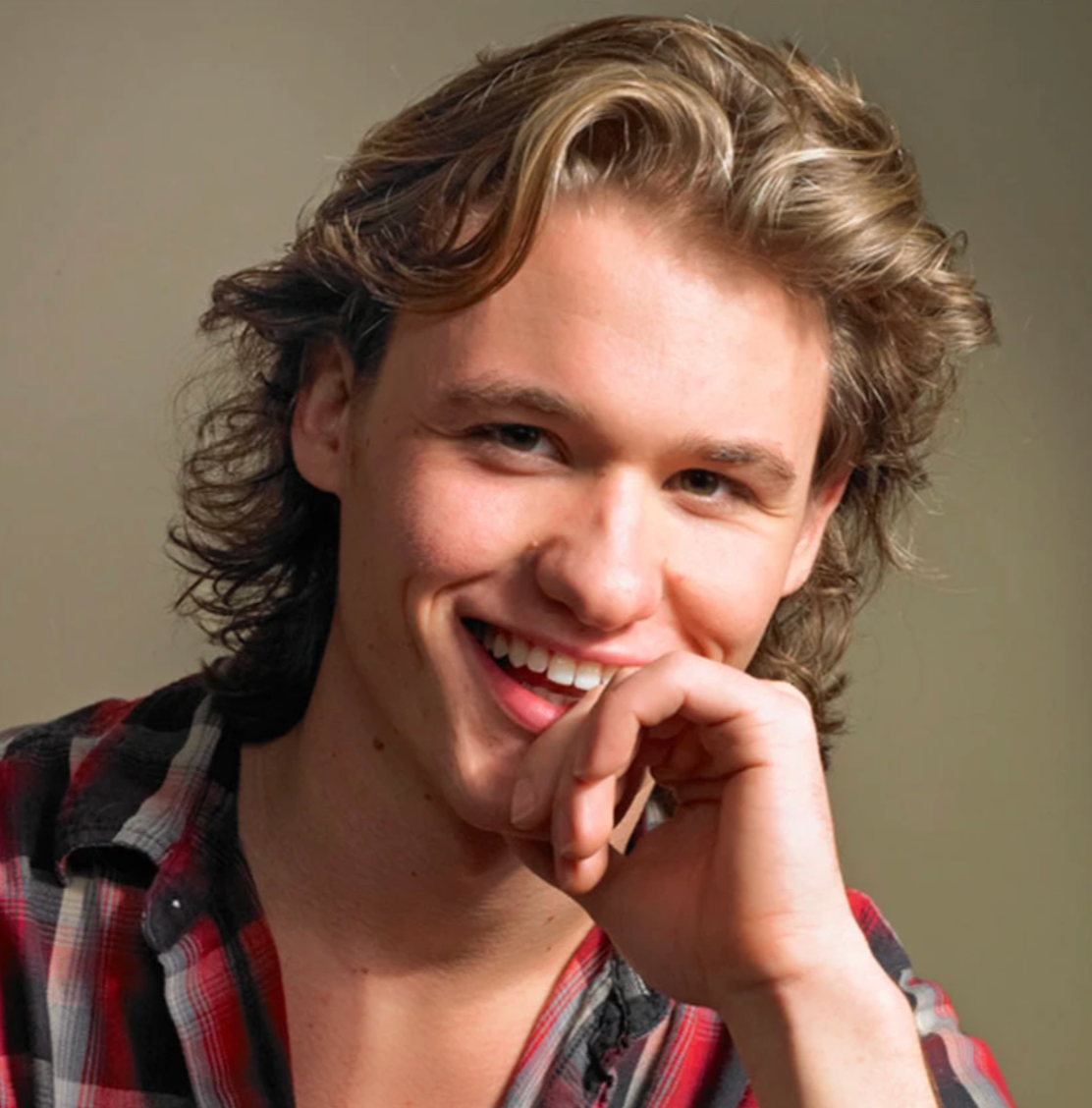 A young man with wavy, light brown hair smiling and resting his chin on his hand, wearing a red and black plaid shirt.