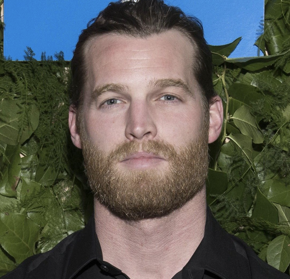 Close-up of a bearded man with slicked-back hair, wearing a black shirt, standing in front of green leaves against a bright blue sky.