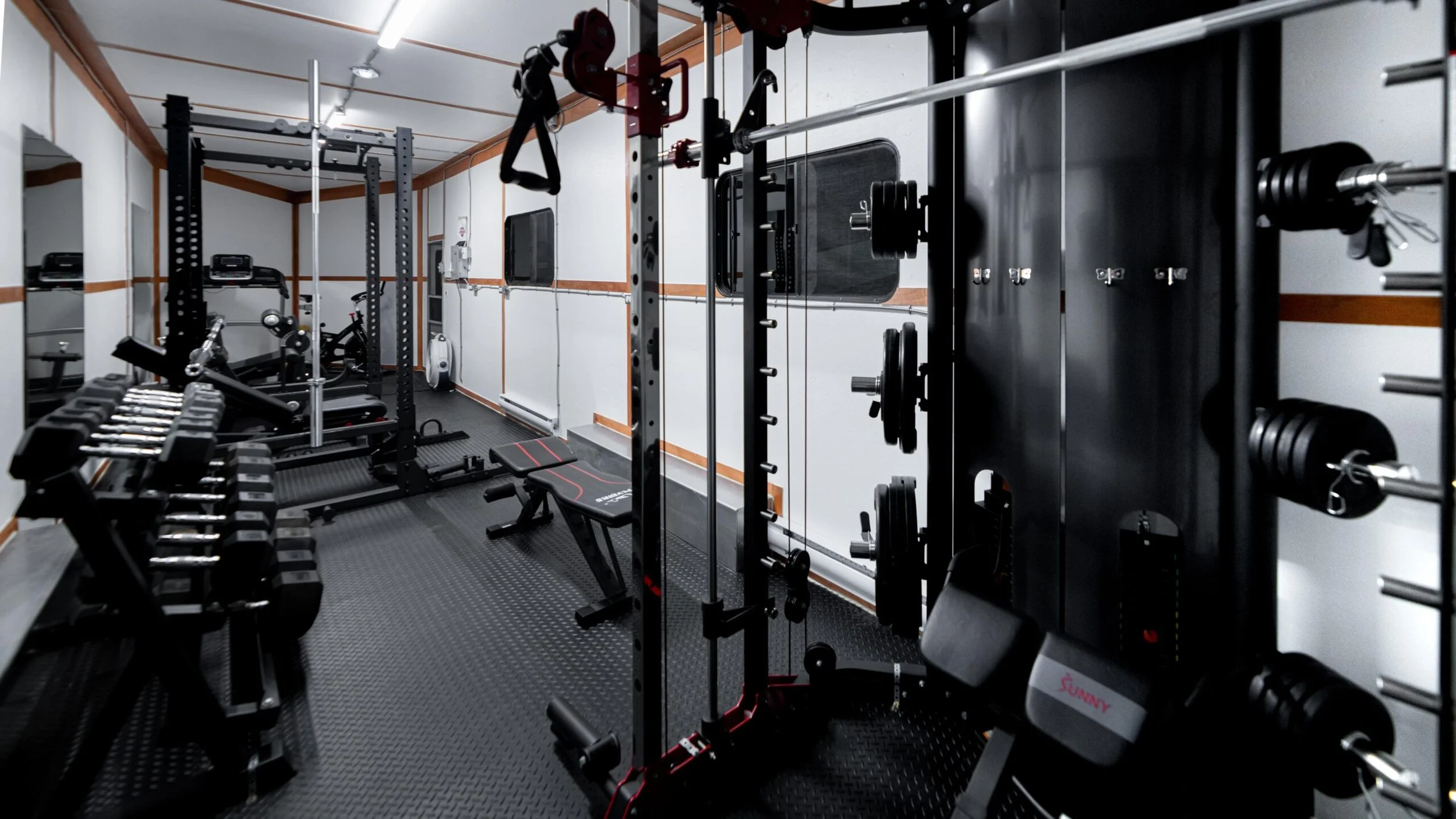 A compact home gym with black and gray workout equipment, including dumbbells, a weight rack, a bench, and a cable machine, situated in a room with white walls and wood trim.