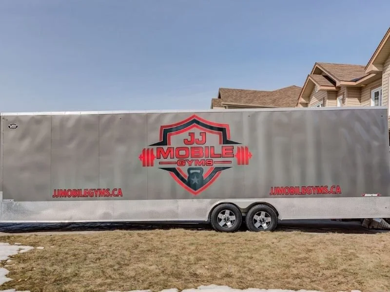 Gray mobile gym trailer with J J Mobile Gyms logo and website, parked on a grassy area in front of residential houses.