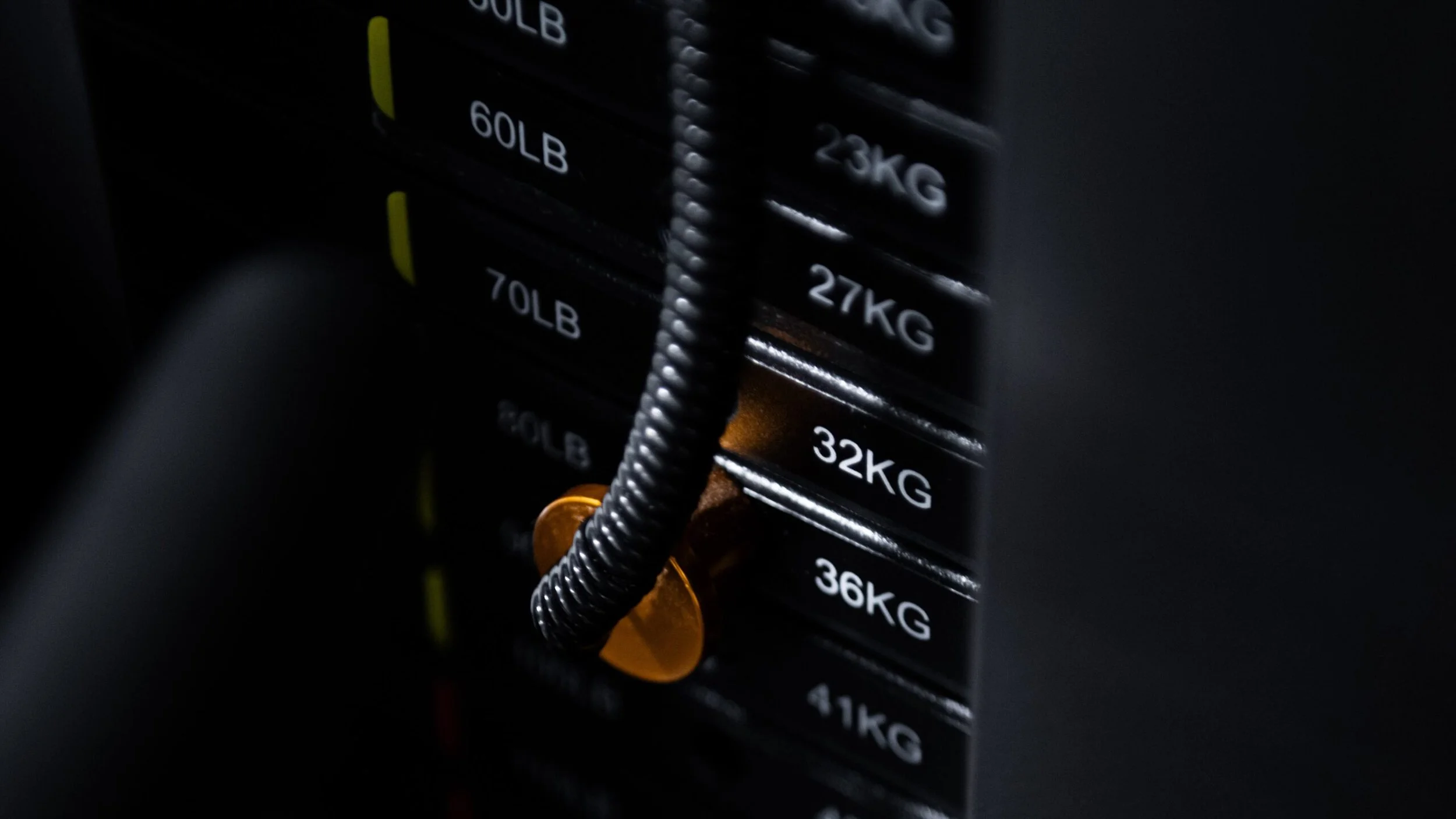 Close-up of a weight plate rack in a gym, showing different weight plates labeled 60LB, 70LB, and 32KG, with one orange weight plate attached to a barbell.
