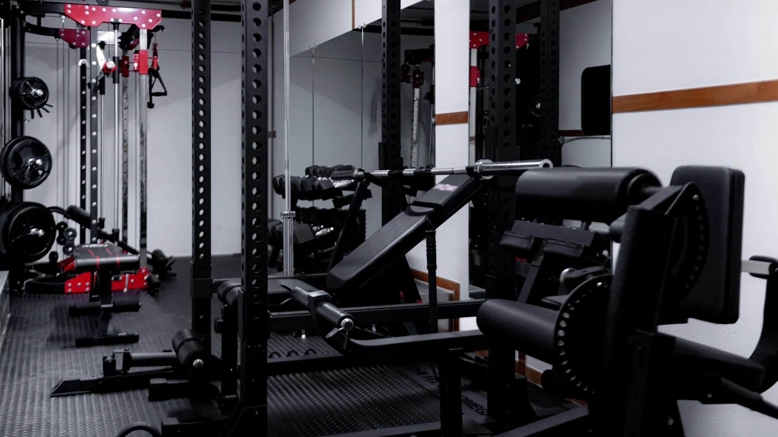 A gym with weightlifting equipment, including a rowing machine in the foreground, and a rack of weights and other fitness gear against the wall.