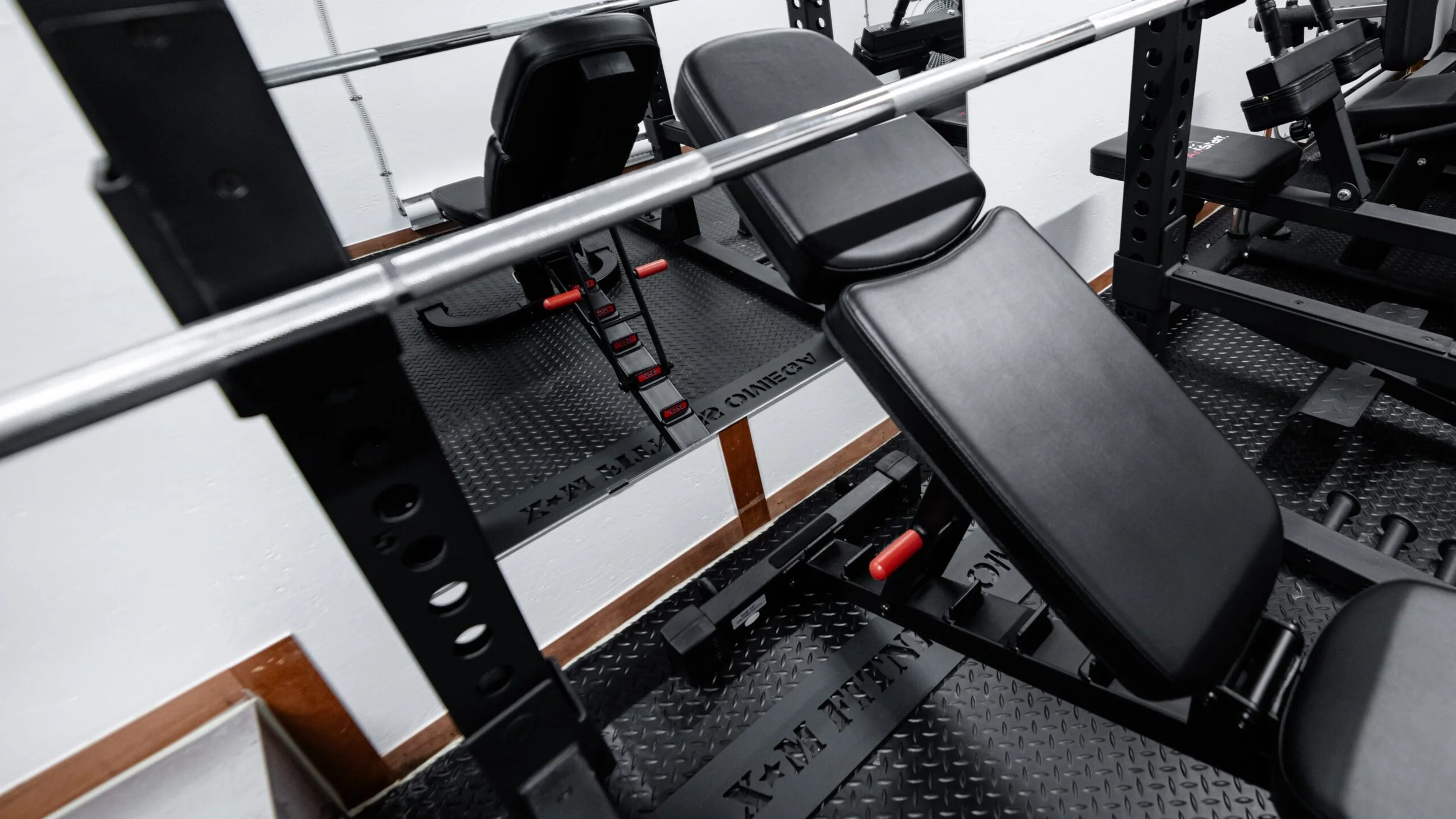 Black adjustable weight bench in a gym next to a power rack with a barbell across the top. Small steps with red handles are underneath the bench. Flat black tiles on the floor with a white wall and wooden baseboard.