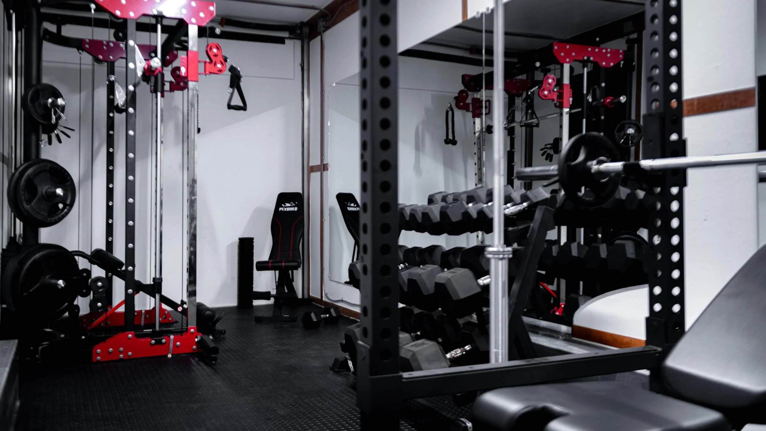 Gym weightlifting area with black and red equipment, including a squat rack, dumbbells, weight plates, and a bench, in a room with black flooring and white walls.