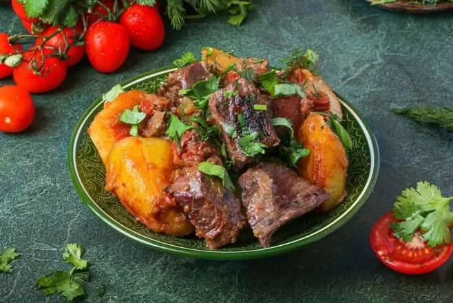 A plate of beef stew with potatoes and carrots, garnished with cilantro, on a green textured surface, with cherry tomatoes and cilantro around it.