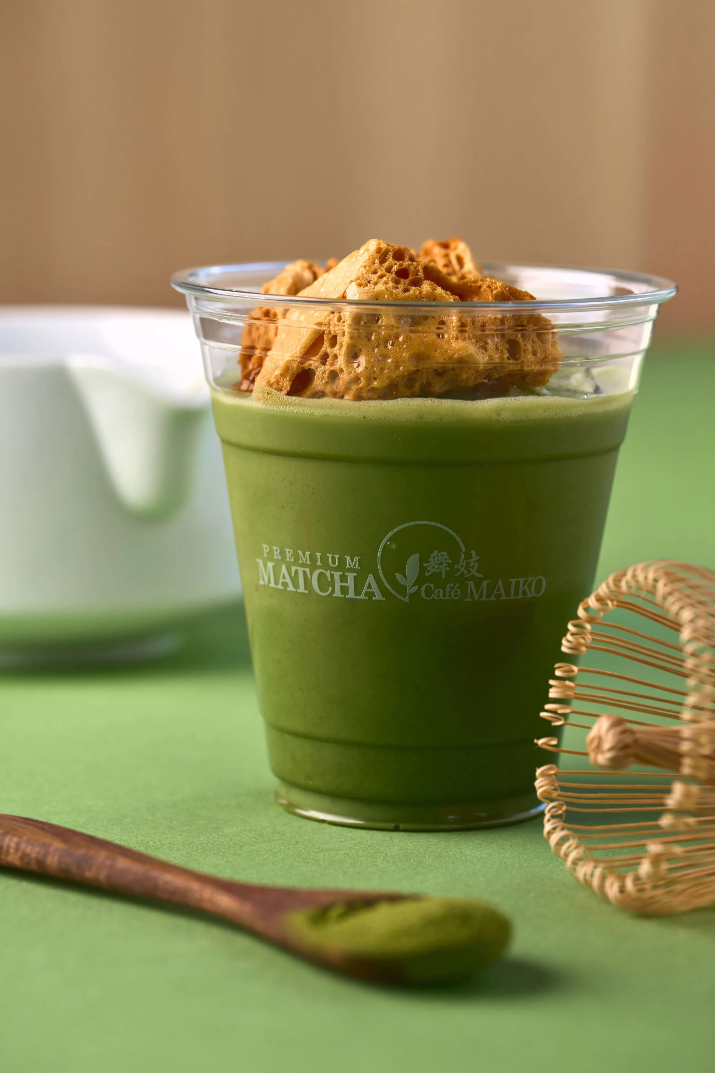 A Honeycomb Matcha Latte on a green surface with a wooden spoon in the foreground and a bamboo whisk on the side.