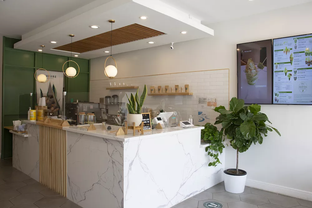Interior of Matcha Cafe Maiko with a white marble counter, green wall, hanging pendant lights, digital screens displaying the menu, and potted plants.