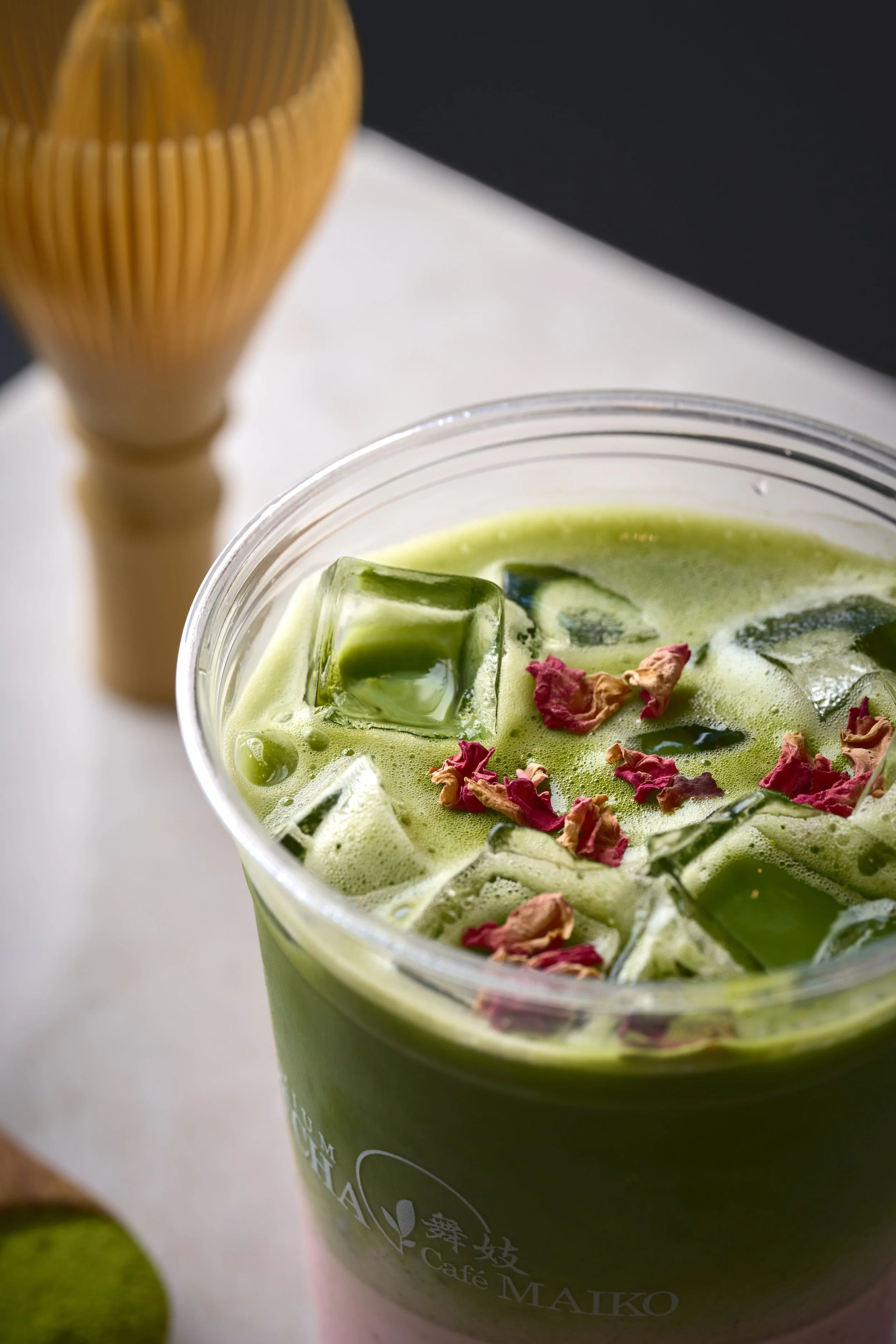 A close-up view of a Matcha Cafe Maiko Rose Matcha Latte, topped with ice cubes and dried rose petals, with a wooden whisk in the background and a lemon to the left.