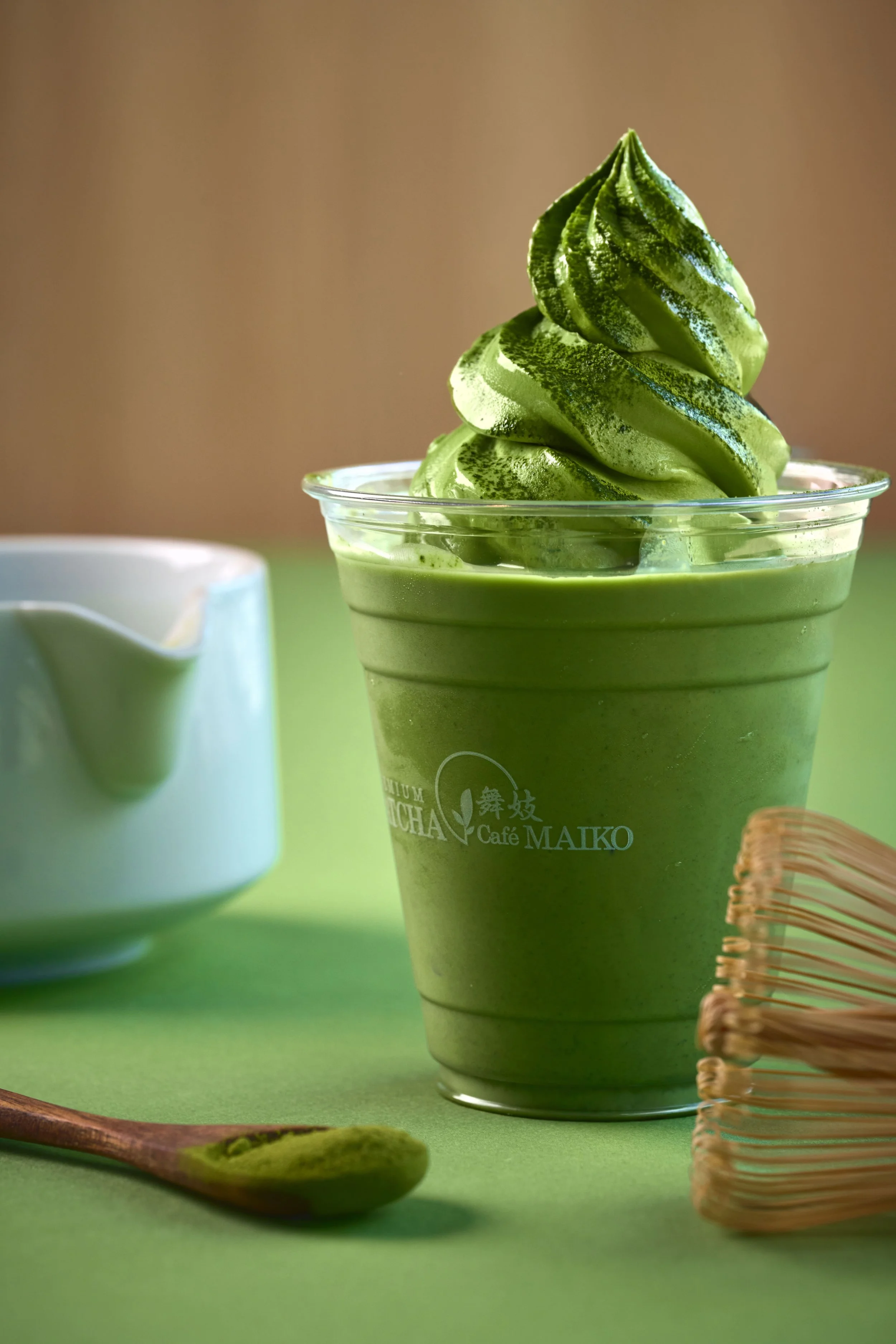 A Matcha Cafe Maiko Matcha latte float, topped with a matcha powder swirl, on a green surface with a wooden spoon and a small woven basket beside it.