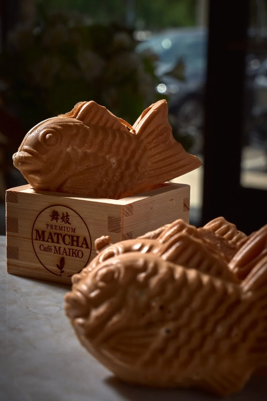 Close-up of fish-shaped pastries called Taiyaki, displayed on a table with sunlight and shadows in the background. One Taiyaki is in a wooden box with Japanese characters and text reading "Premium Matcha Cafe Maiko."